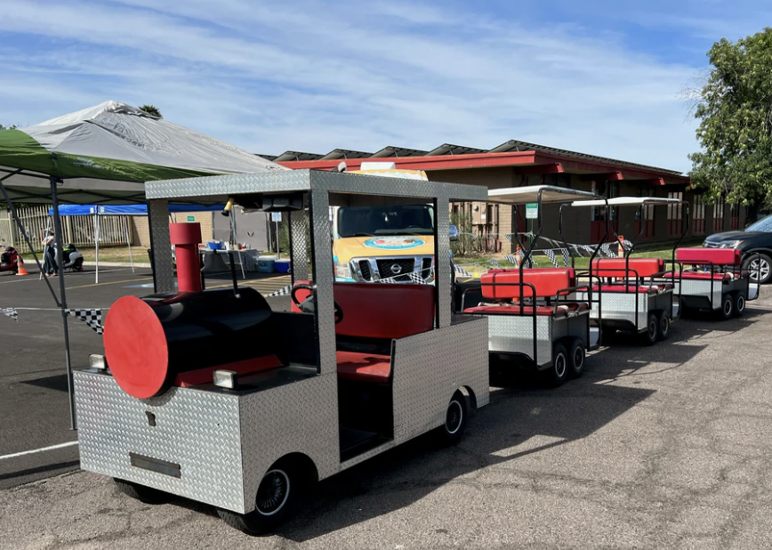 Small train ride vehicles with a train in front, parked outdoors near tents and a building, under a blue sky