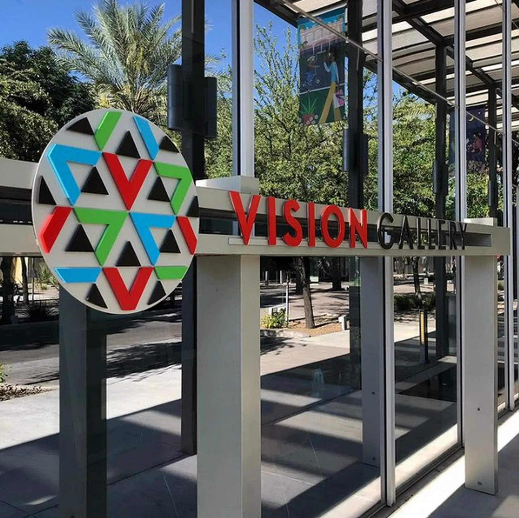 Close-up of a modern glass entrance to a building with a sign that reads 'VISION GALLERY' in red letters. Behind the glass, there is a colorful poster and some trees with green foliage and a clear blue sky.