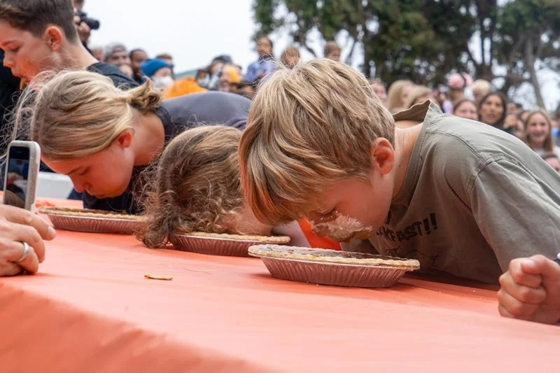 Children participating in a pie-eating contest, with their faces in pies, while a crowd watches in the background.