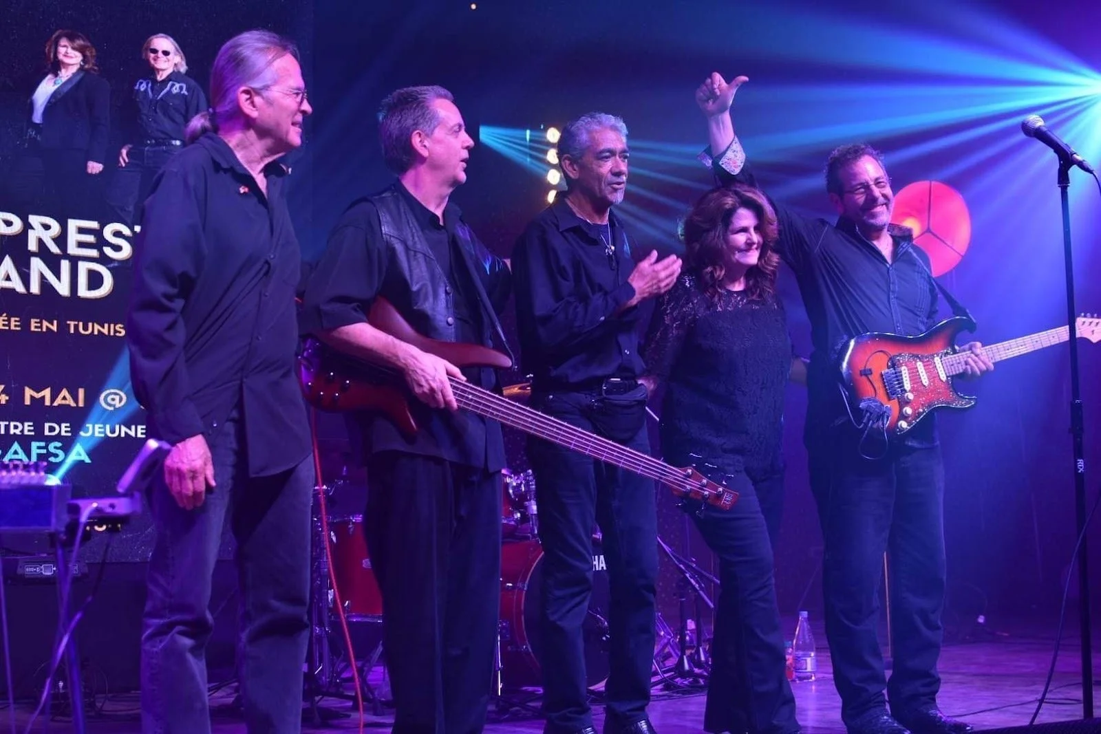 Five musicians with guitars on stage during a concert, some clapping and one raising an arm, with stage lights and a colorful backdrop in the background.