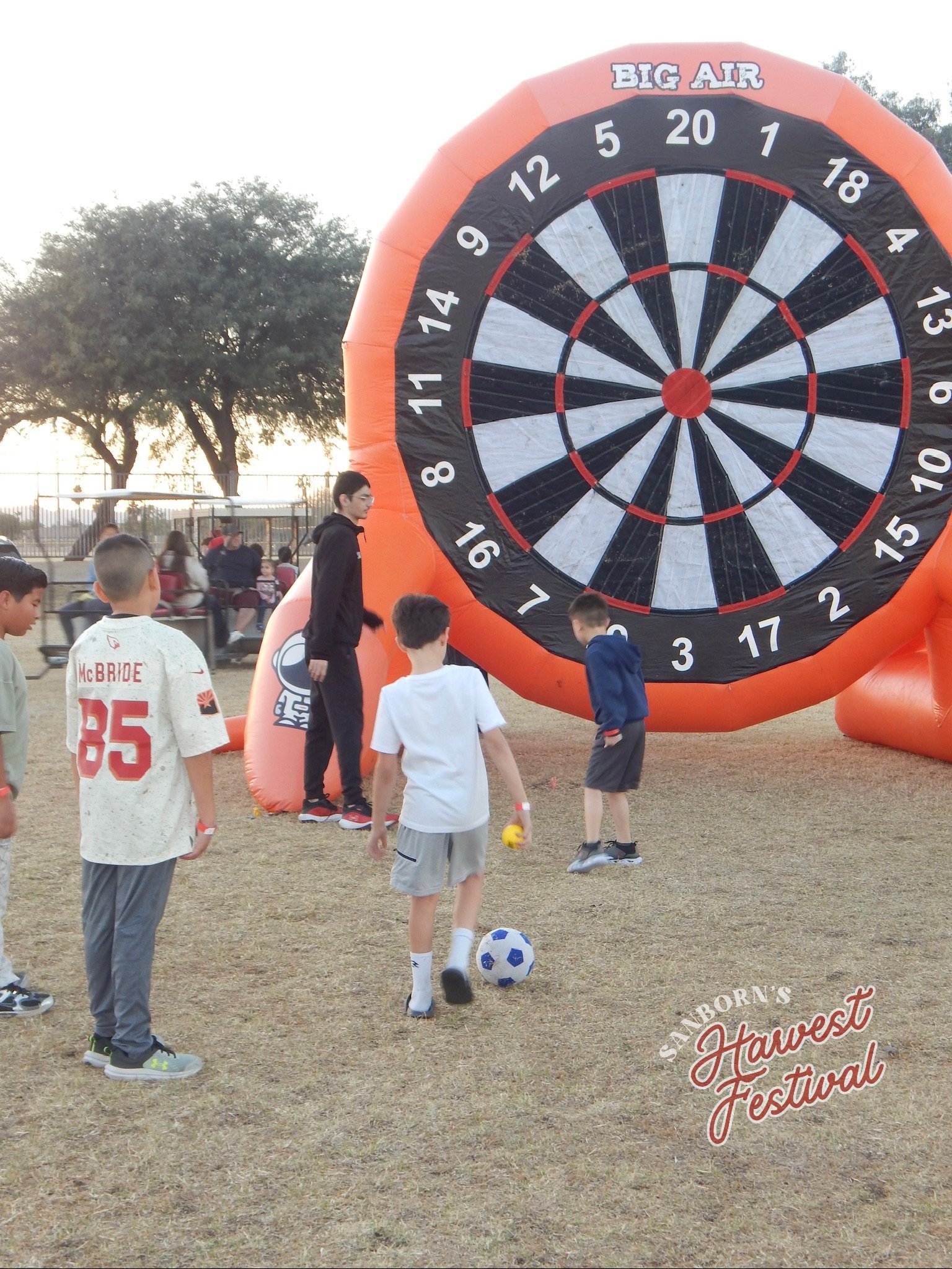 Children playing soccer in front of a large inflatable dartboard at a festival. Some children are kicking soccer balls, and others are standing and observing. Trees and a fence are visible in the background, and the event appears to be outdoors durin