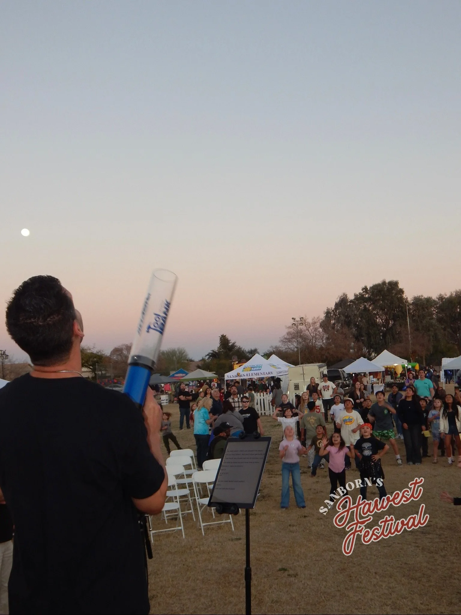 A crowd at an outdoor festival with tents, trees, and a clear sky at sunset. The festival is called 'Sanborn Harvest Festival' and people are gathered around, some looking towards the sky.