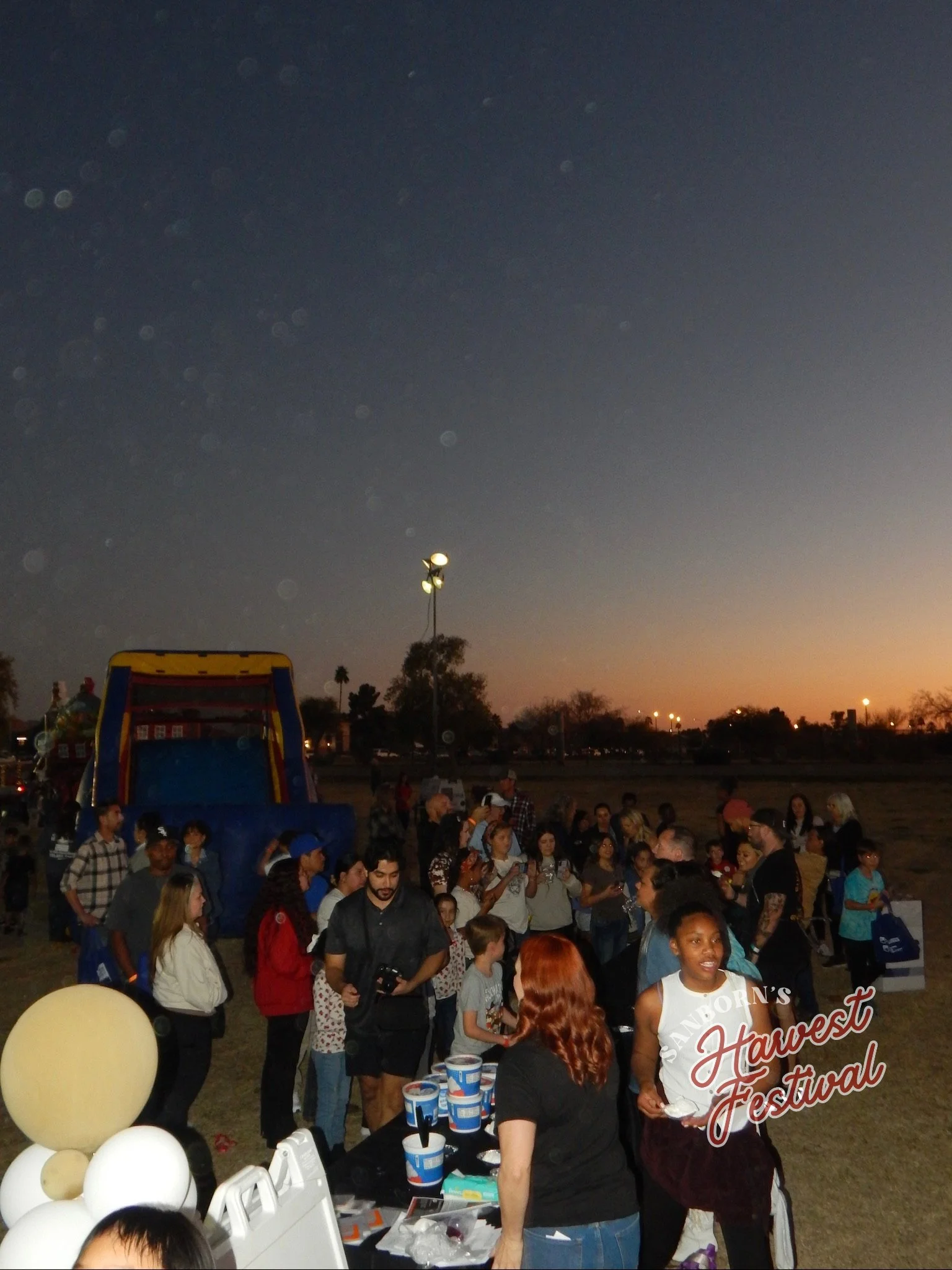 A crowded outdoor festival during dusk, with many people gathered around a booth serving food and drinks. There are balloons and a large inflatable bounce house in the background, with a darkening sky and some lights visible in the distance.
