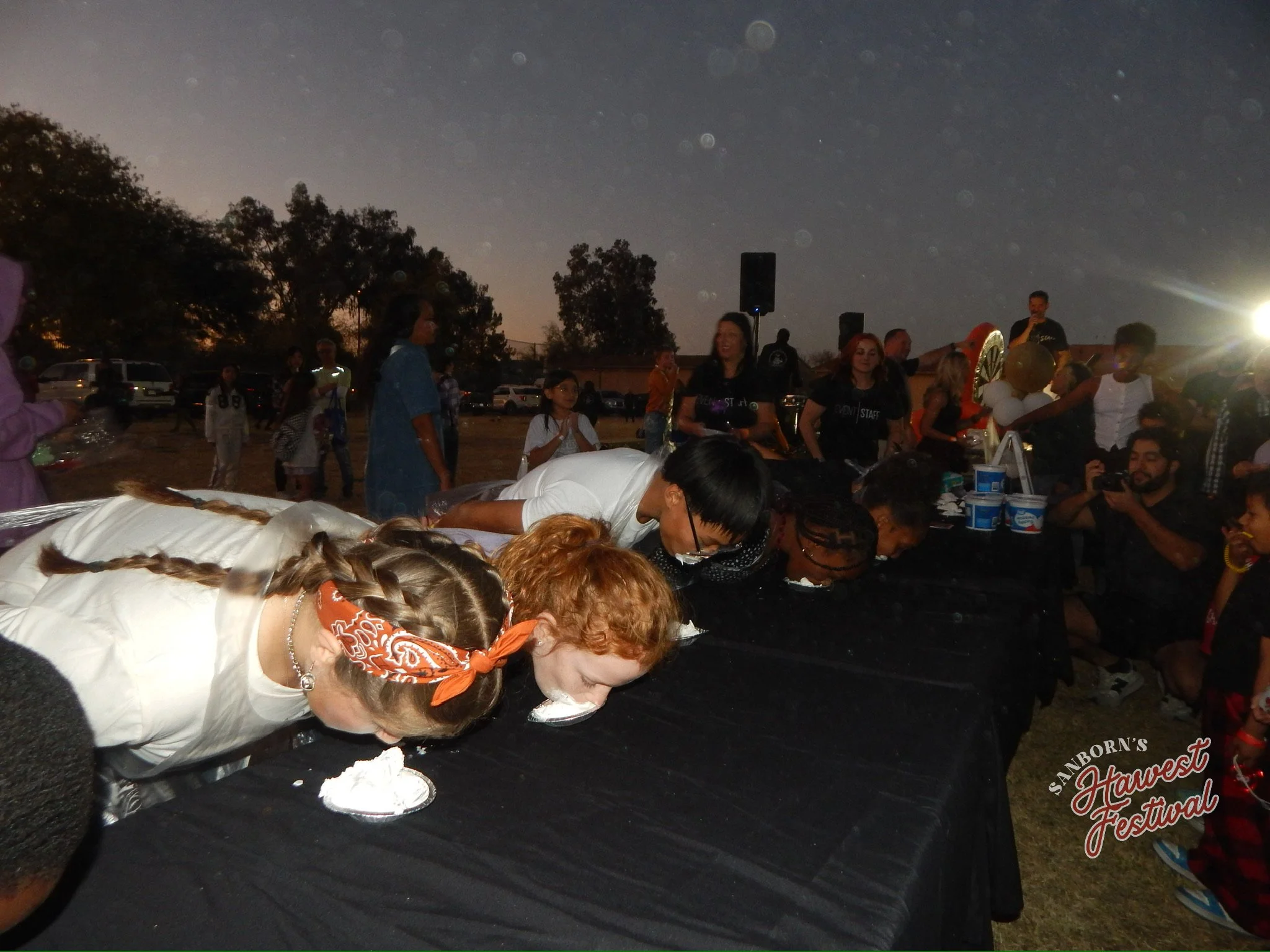 People bowing their heads during an outdoor event at sunset with trees and cars in the background, at the Sanborn's Harvest Festival.