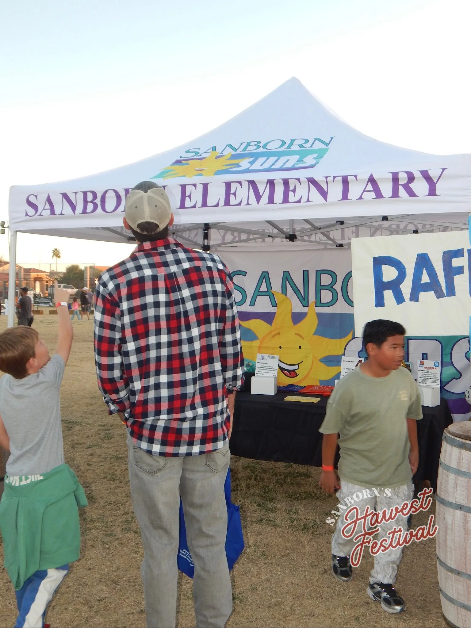 People at Sanborn Elementary School booth at a fair or festival, with children and adults near tents and displays outdoors.
