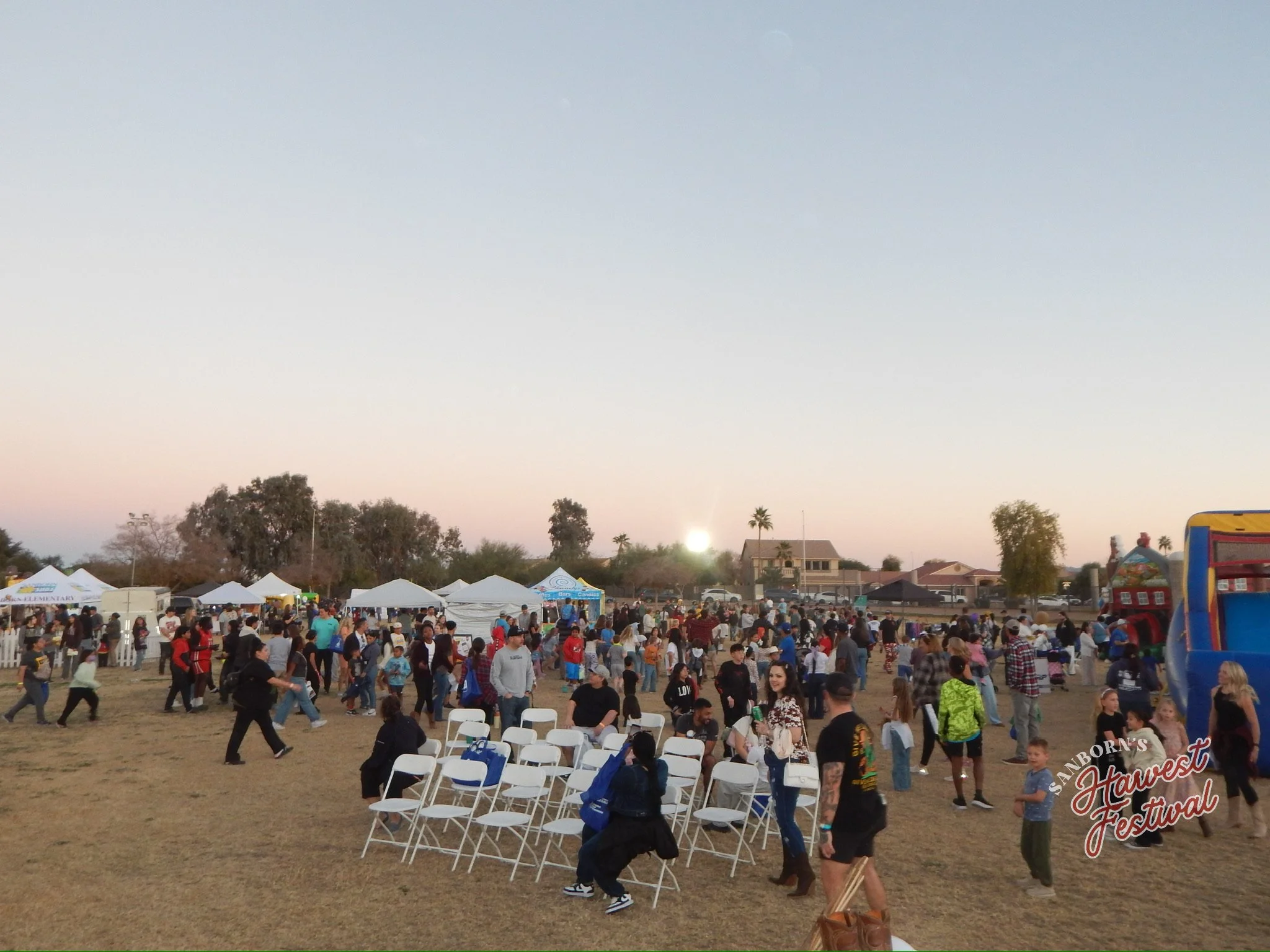 People attending a festival outdoors with tents, trees, and a few buildings in the background during sunset.