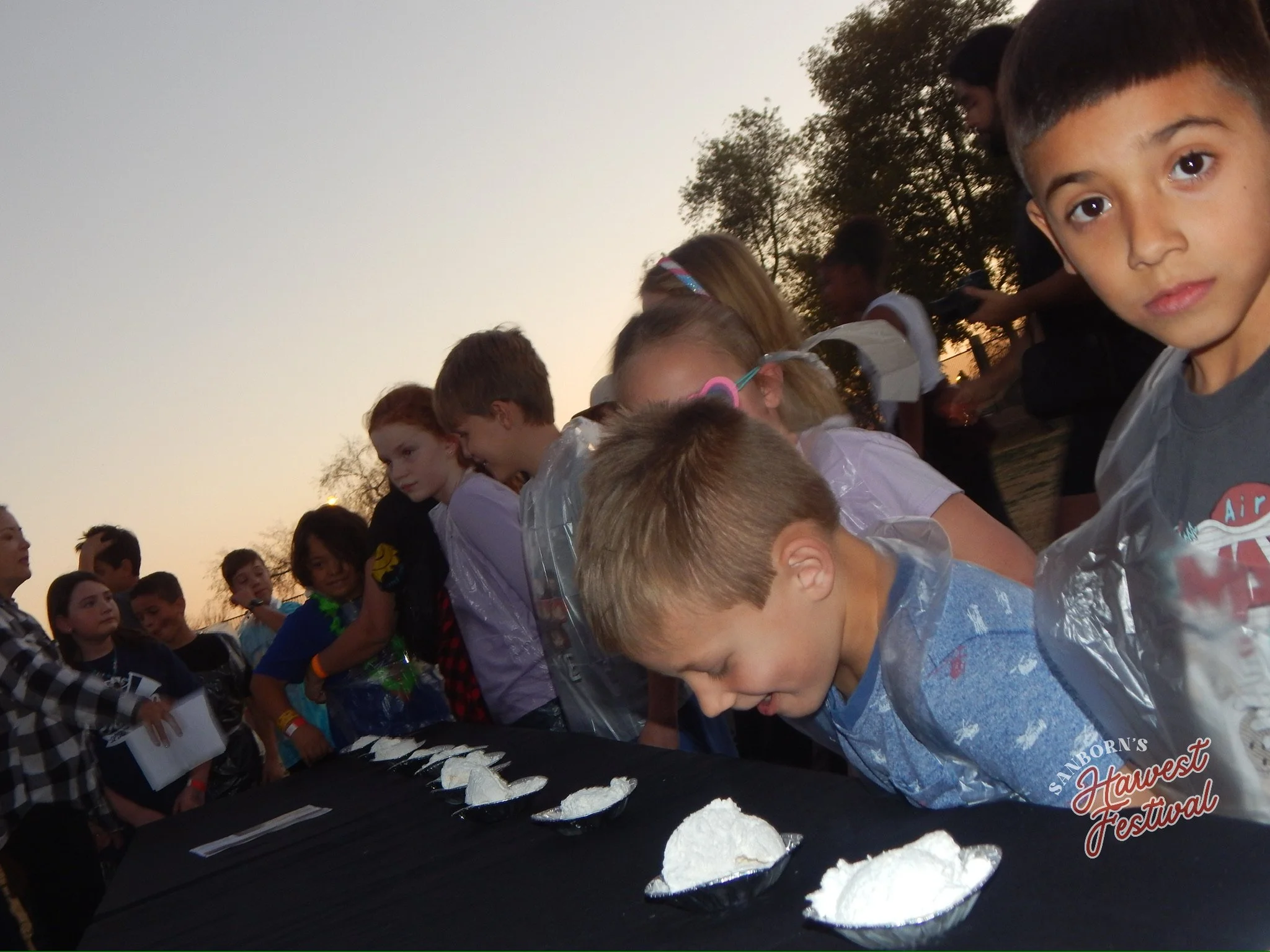 Children gathering around a table at an outdoor event during sunset, with some children wearing plastic ponchos and others with accessories like glasses, while a cake with white frosting and pieces is on the table, part of a festival celebration.