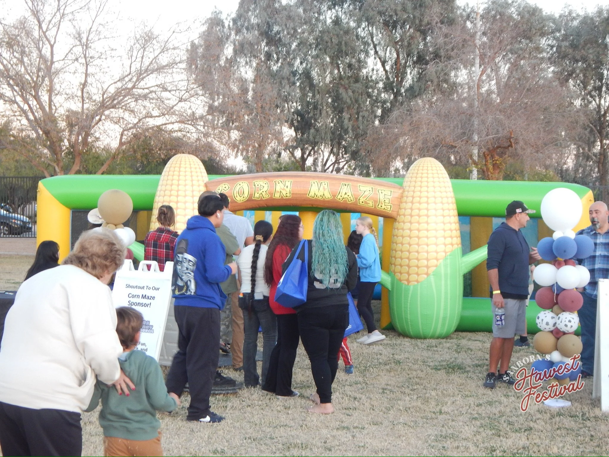 At an outdoor festival, a vibrant inflatable corn maze titled 'Corn Maze' is set up, featuring large yellow corn cob decorations at its entrance. Crowd of people, including adults and children, gather around, some engaging with the maze and others so