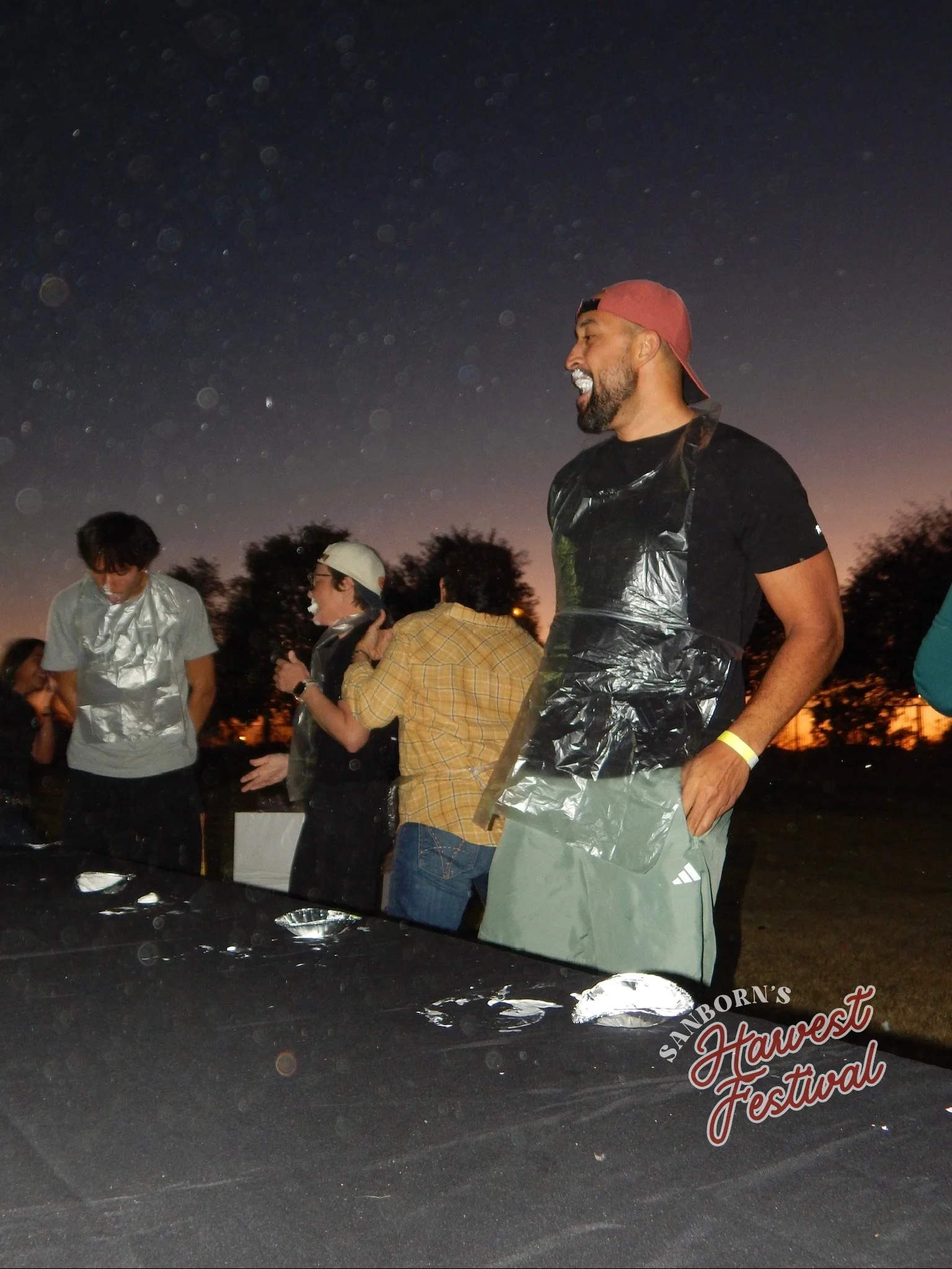 People gathered at an outdoor event during sunset, with a man smiling and wearing a red cap and plastic apron, possibly carving or preparing food, at the Sandborn's Harvest Festival.