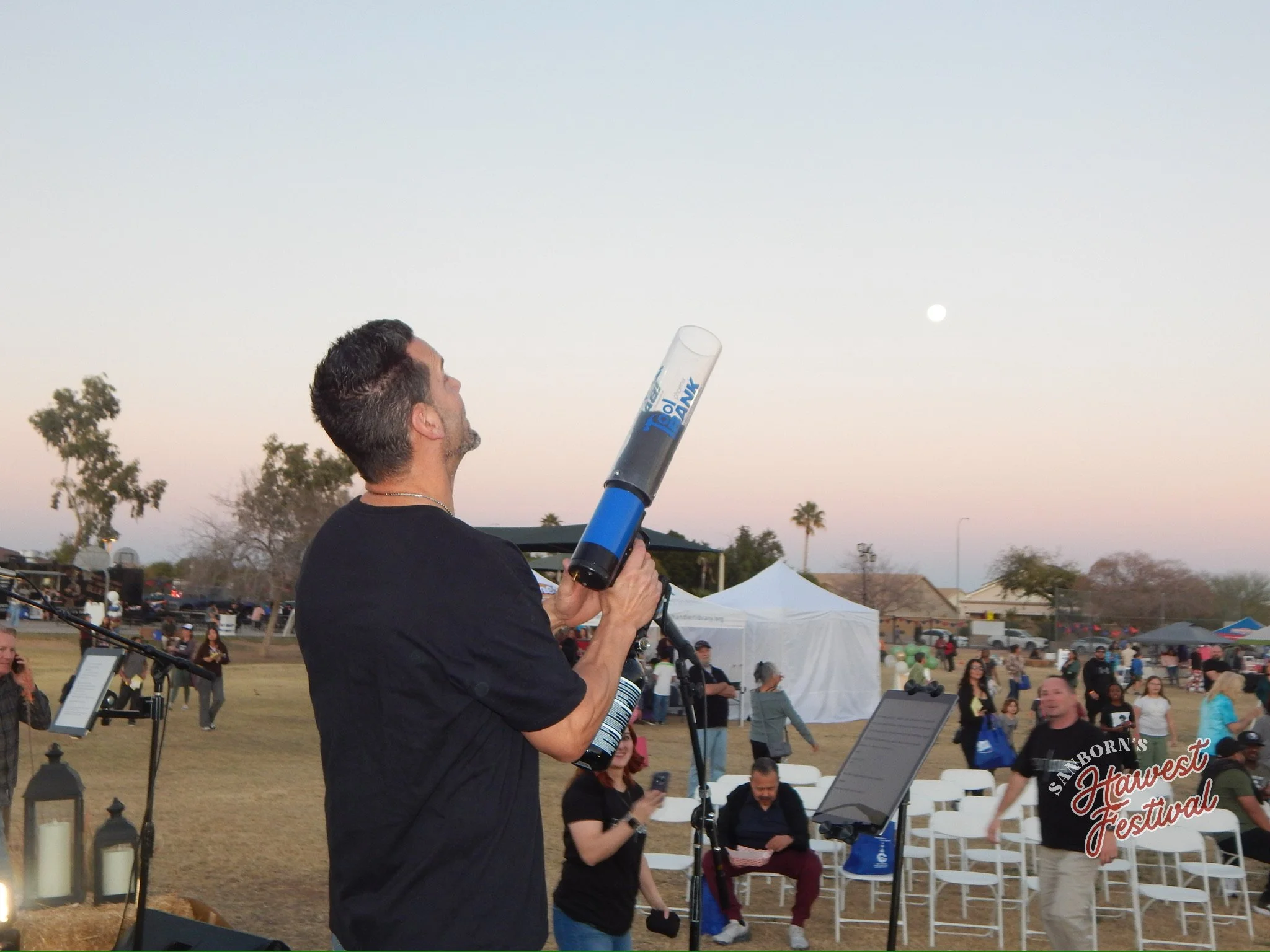 A man with short dark hair and beard holding a tube of fireworks or a rocket at an outdoor event during dusk, with a crowd of people, tents, and trees in the background.