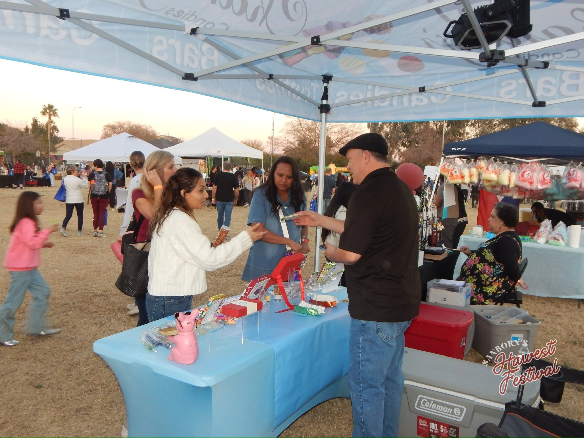 People at an outdoor festival booth sharing and purchasing small toys and gifts, with other vendors and festival activities visible in the background.