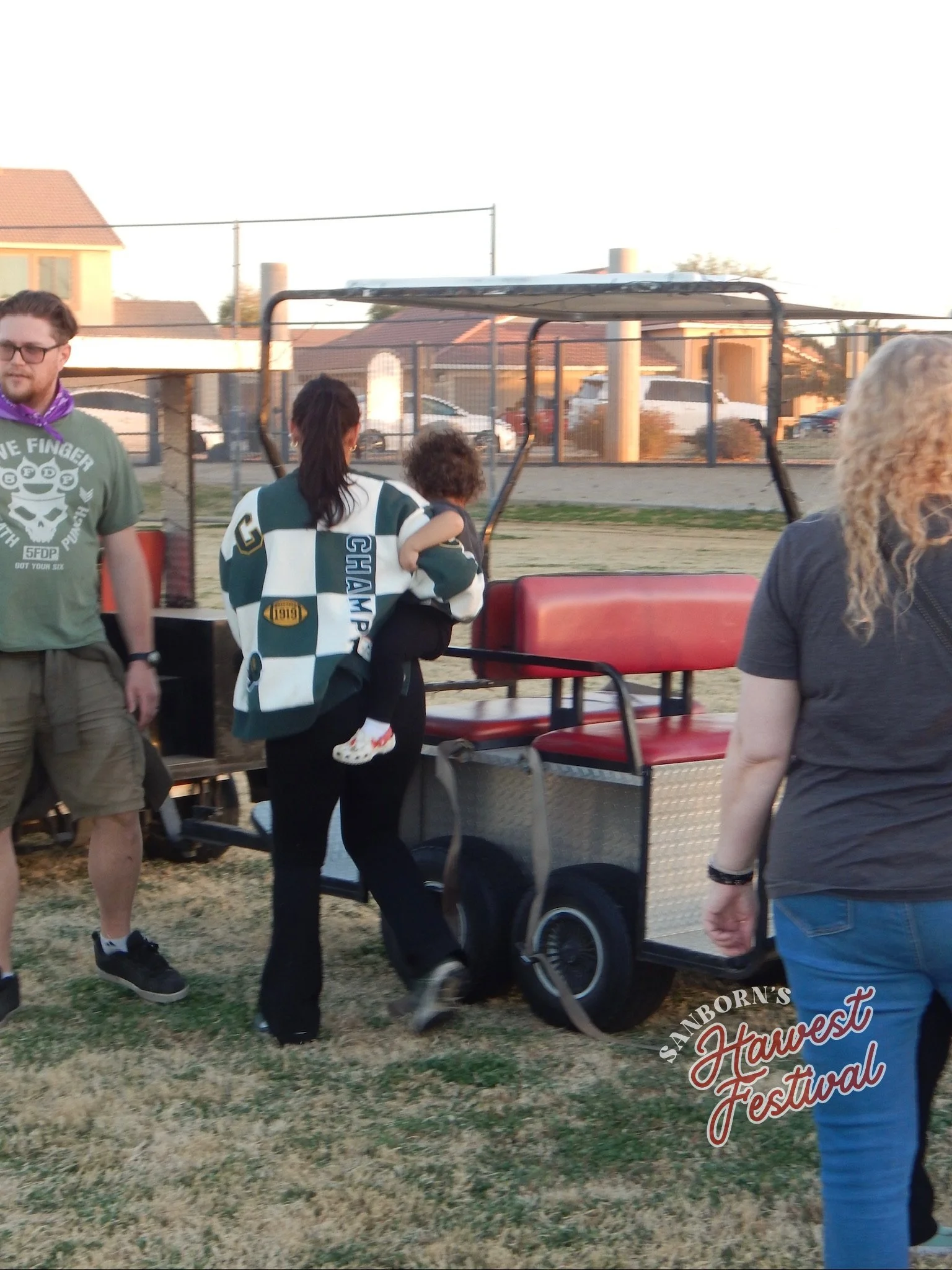 People standing outdoors at a festival, including a woman holding a child, with a sign for Sanford's Harvest Festival visible in the foreground.