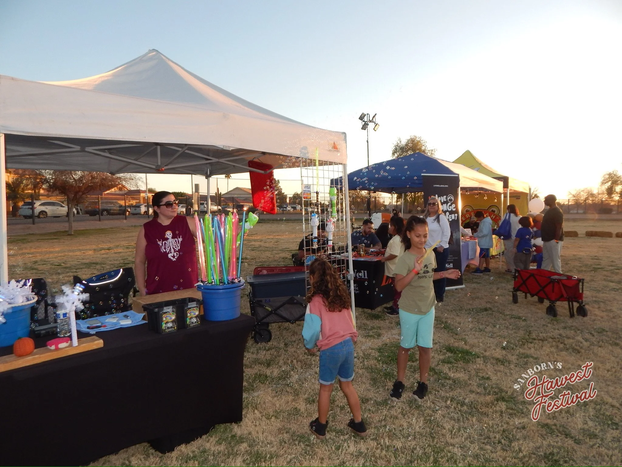 People at an outdoor festival with tents, vending booths, and children. The sky is clear with the setting sun, and there are trees and parked cars in the background.