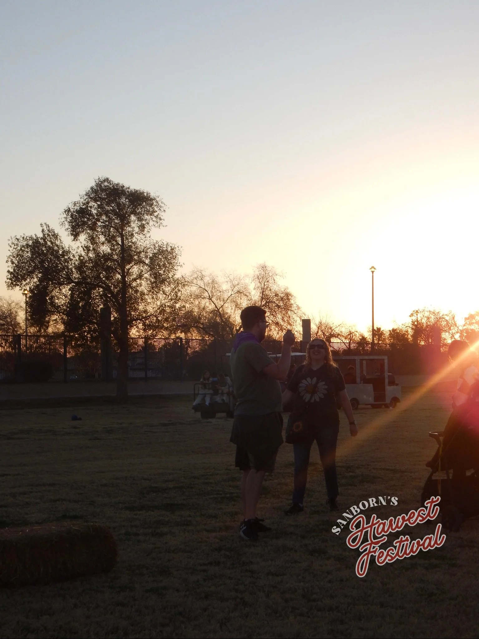People at an outdoor harvest festival during sunset, with trees, a fence, and festival signage in the background.