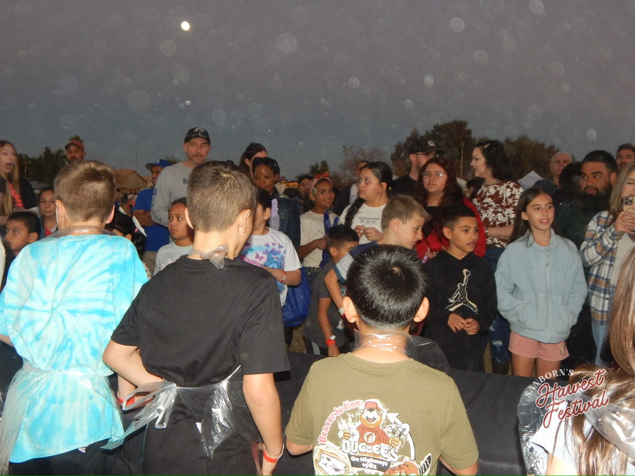 A group of people, including children and adults, at a festival outdoors at night, with a visible full moon in the sky.