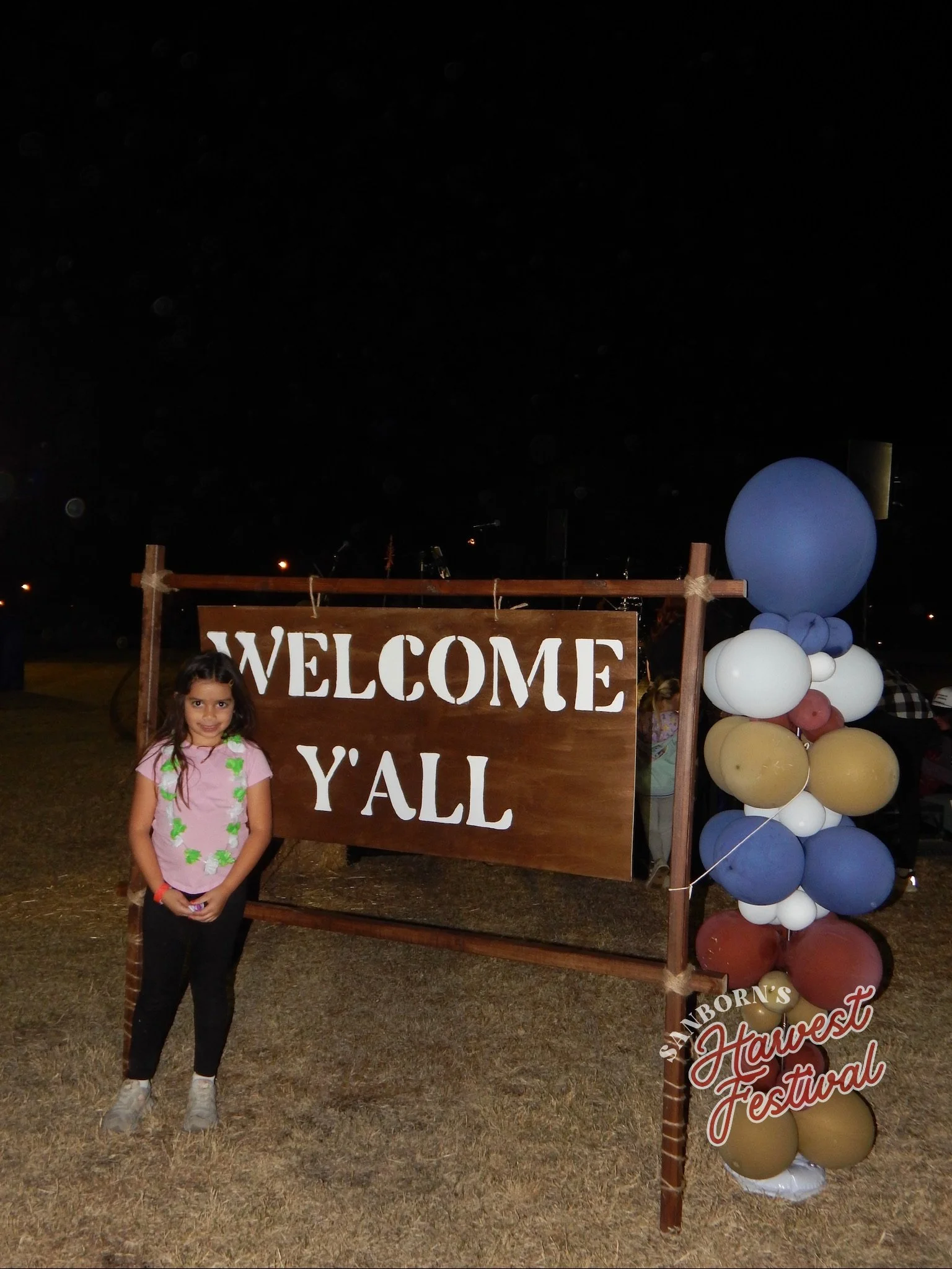 A young girl standing in front of an outdoor welcome sign at a fall festival, with balloons on a stand to the right. The sign reads 'WELCOME Y'ALL,' with the rest cut off in the photo.