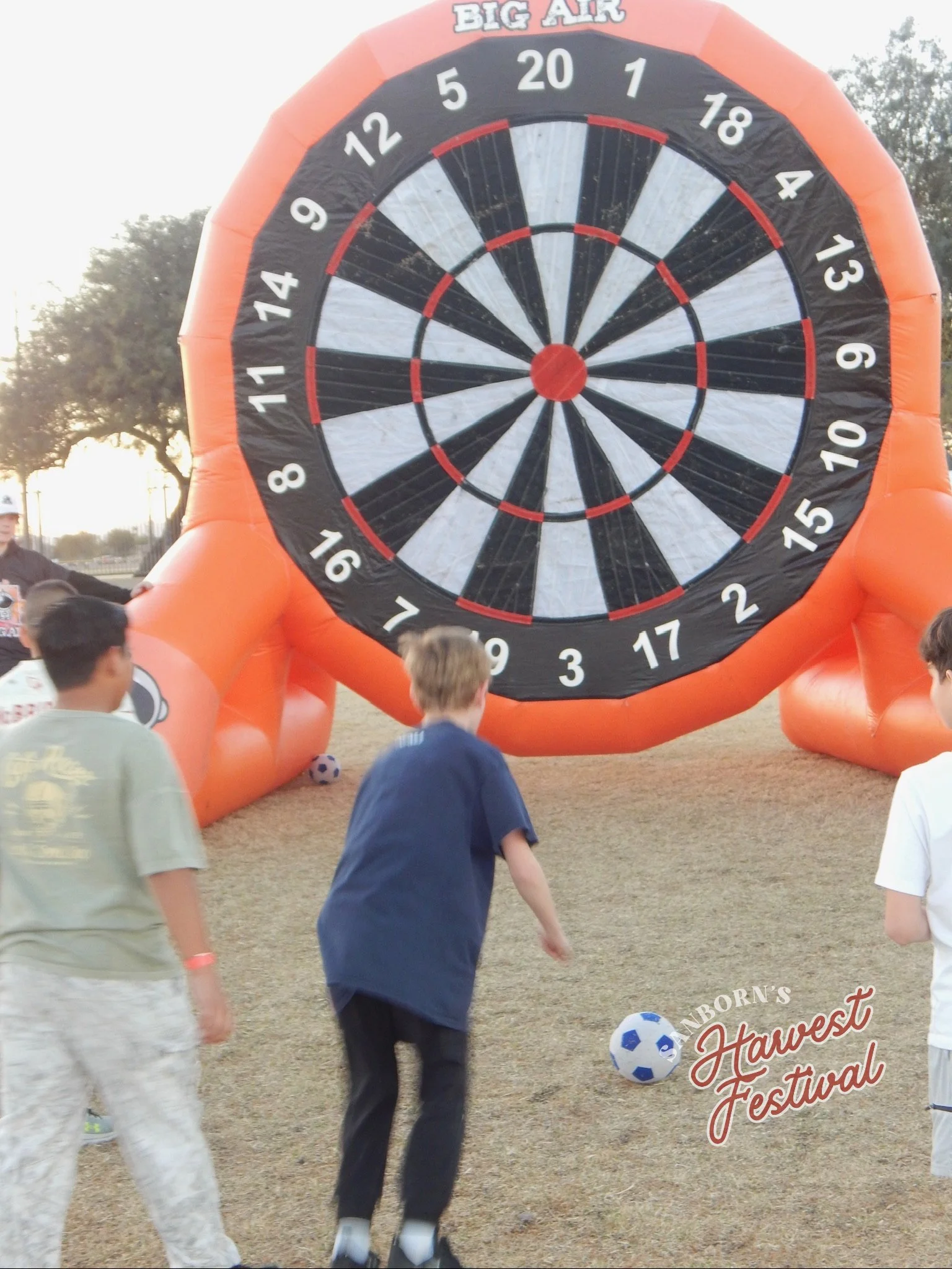 Children playing soccer near a large inflatable dartboard at a fall festival.