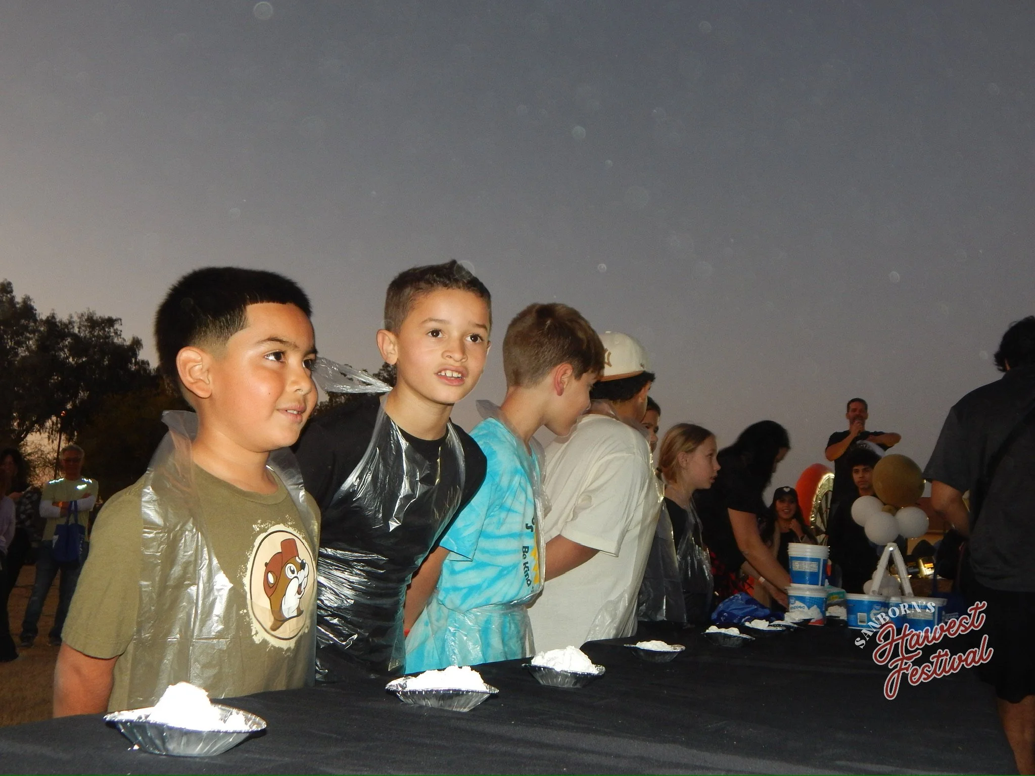 Kids in line at a festival, some wearing plastic rain ponchos, with a table in front of them containing ice cream or frozen treats, and a crowd of people in the background under a dark sky.