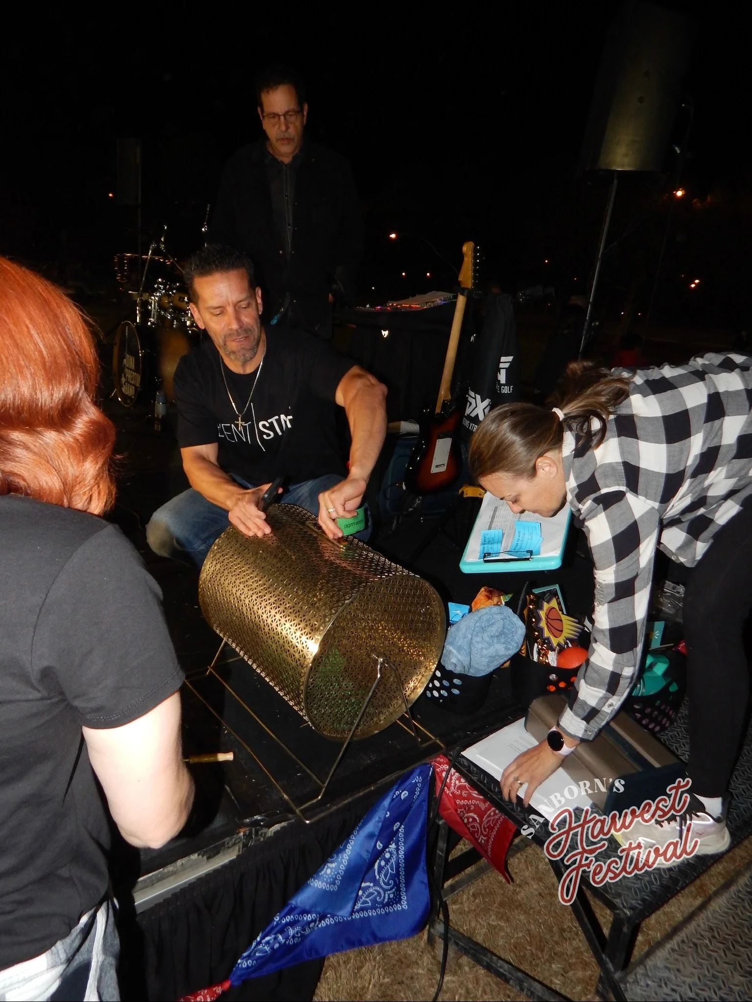 People gathered at a night festival, with a sign reading 'Southwest Festival'. Some are handling tickets or flyers, and there are musical instruments visible in the background.