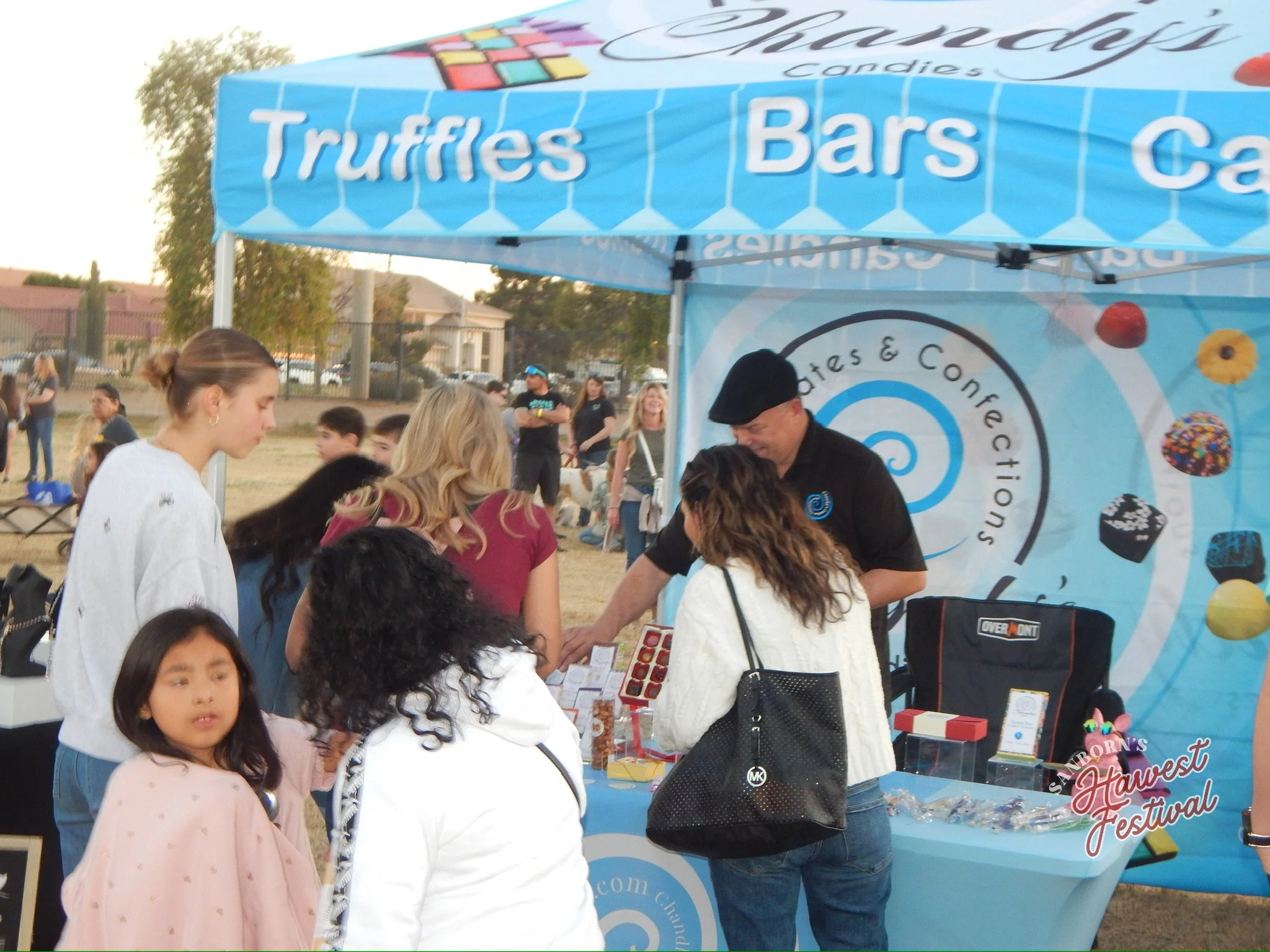 People gathered at a booth during an outdoor festival, with a sign that reads 'Truffles,' 'Bars,' and 'Candy.' There are children and adults engaging with vendors, and the booth appears to sell chocolates and confections.
