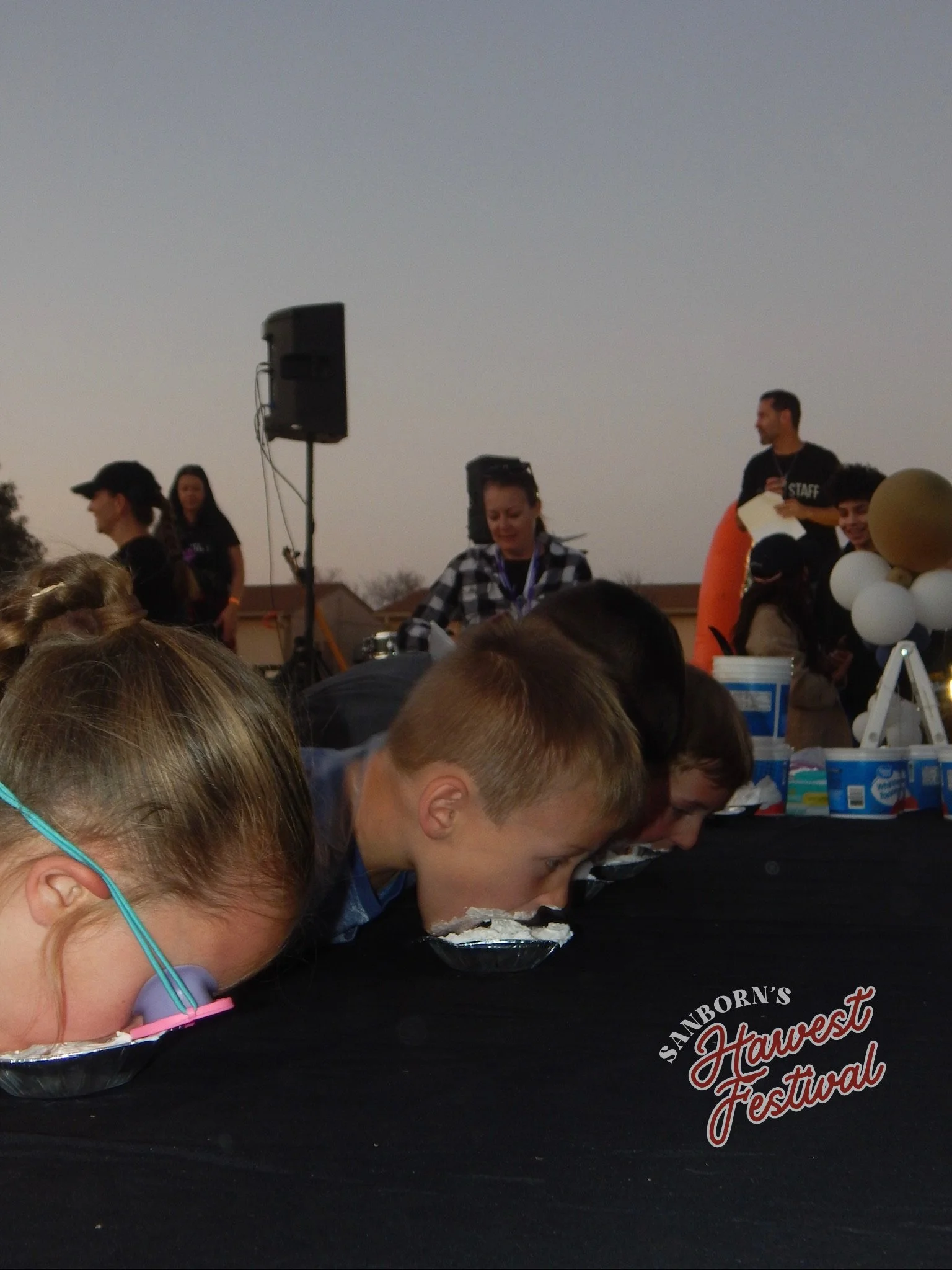 People participating in a pie-eating contest at a harvest festival, with a group of individuals leaning over tables and focused on eating. A man in a black shirt labeled 'STAFF' stands in the background, and balloons and a speaker are also visible.