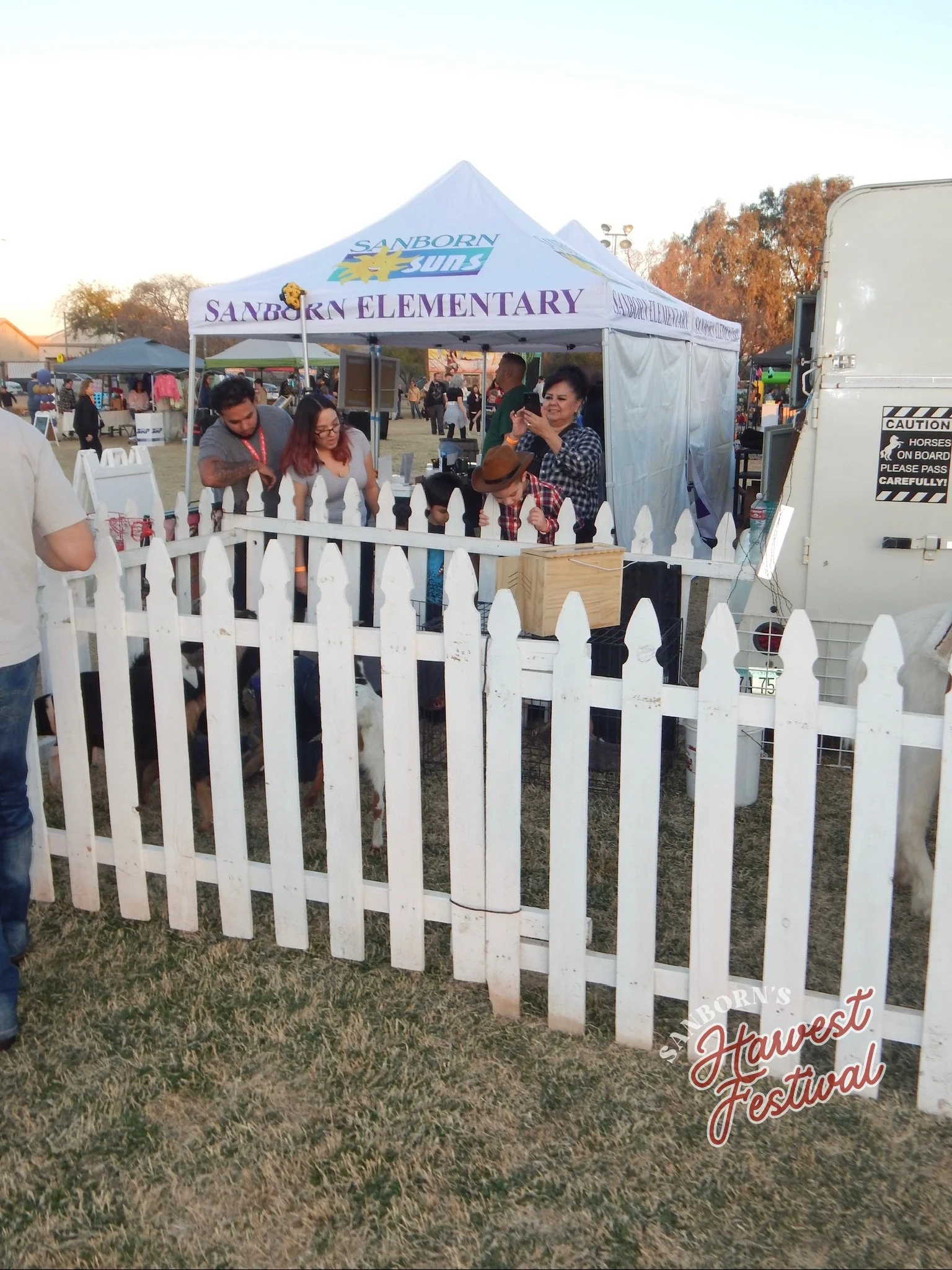 at an outdoor festival, a white tent with purple text reading 'Sanborn Elementary' has people gathered inside, some taking photos or looking at something, with a small fenced area in front. The background shows other tents, trees, and a clear sky.