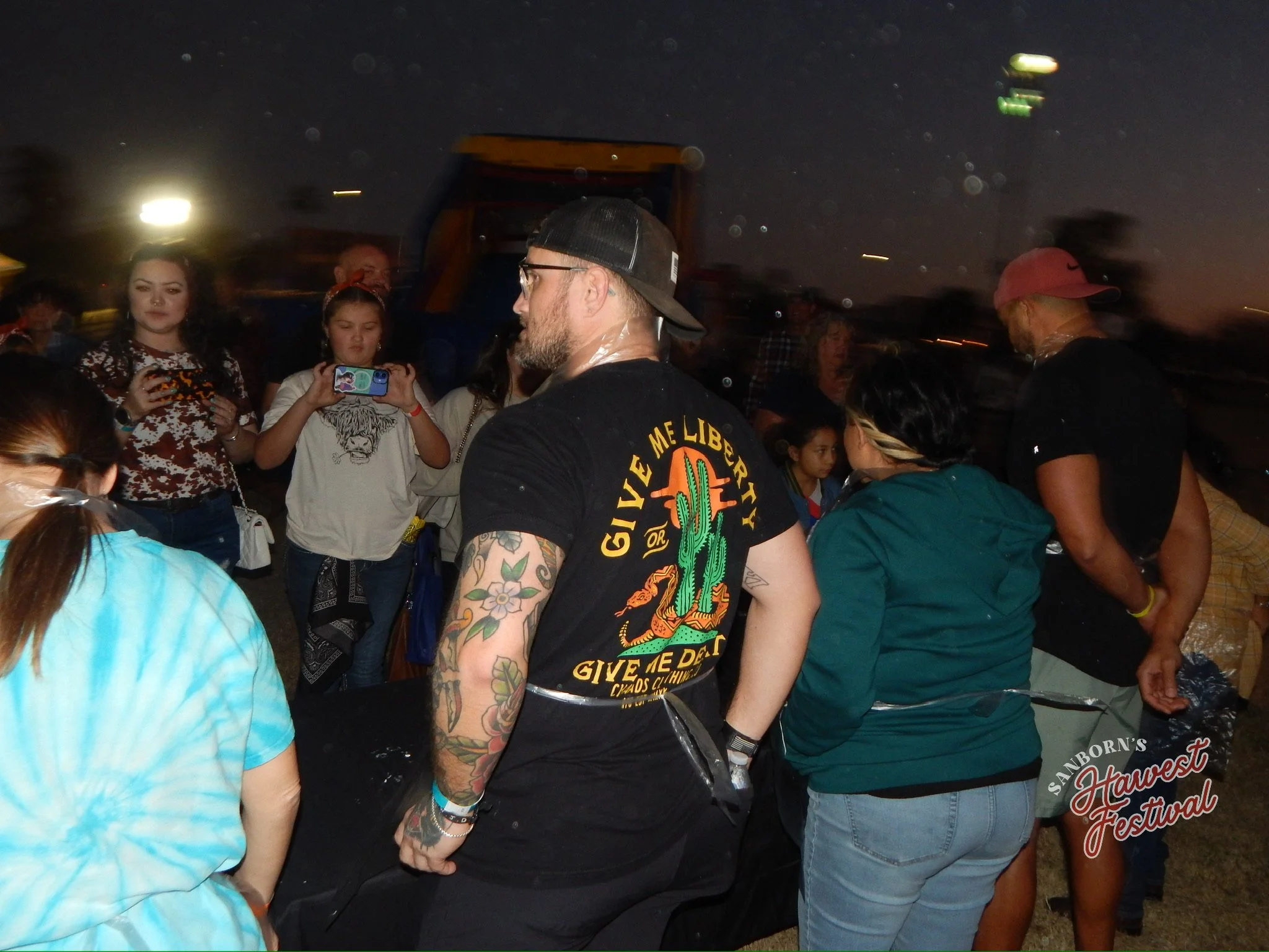 Group of people at a nighttime outdoor event, including a woman with dark hair wearing a cow print shirt, a girl taking a photo, and a man with a beard and glasses wearing a baseball cap and a black T-shirt. The background is dark with some lights an