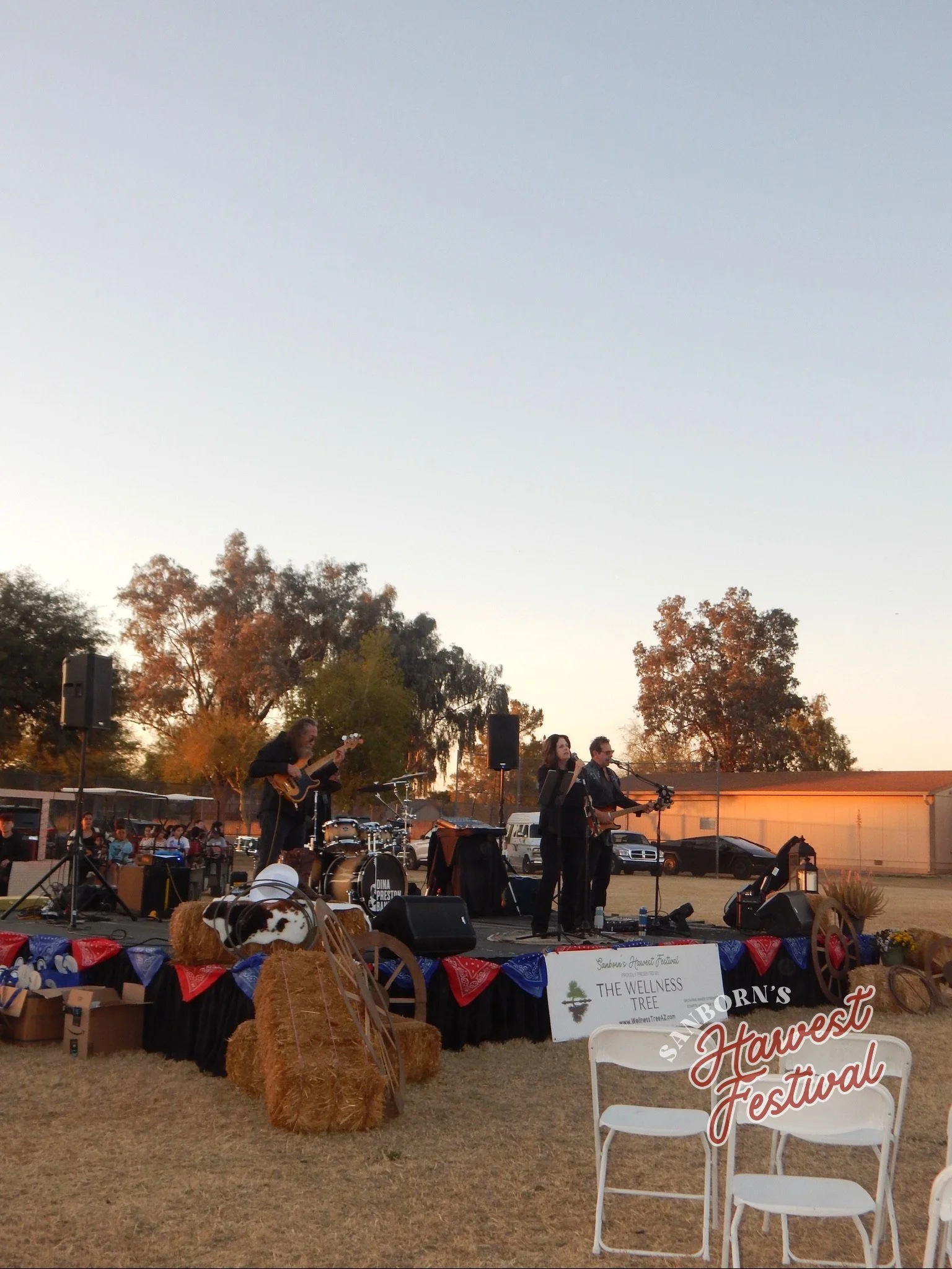 Live outdoor music performance at a local festival with a band playing on stage, surrounded by trees with fall foliage, hay bales, and festival decorations including a sign for the Wellness Tree and Harvest Festival.