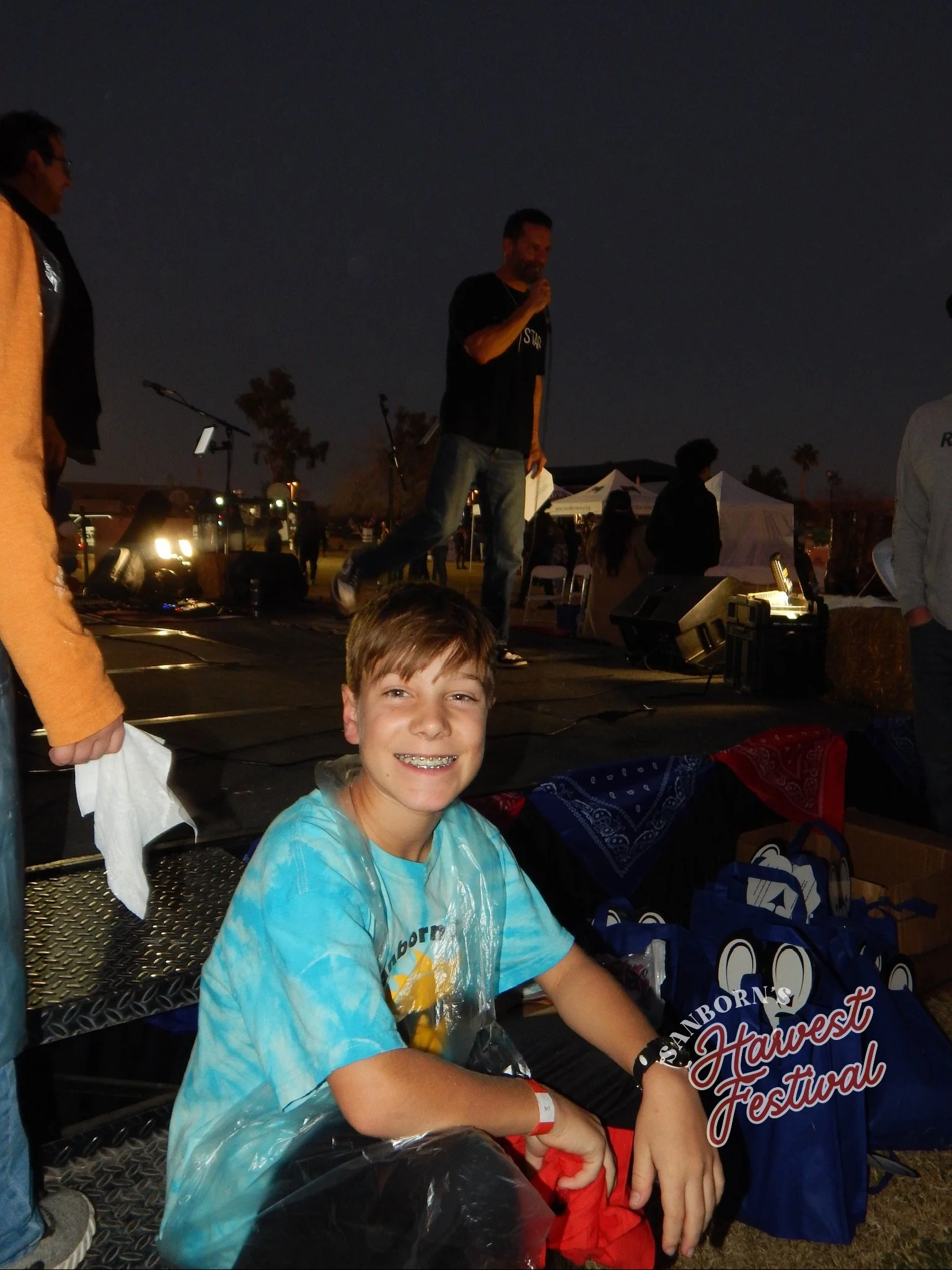 Young boy smiling at the camera sitting at a table during a night festival, with people and tents in the background.