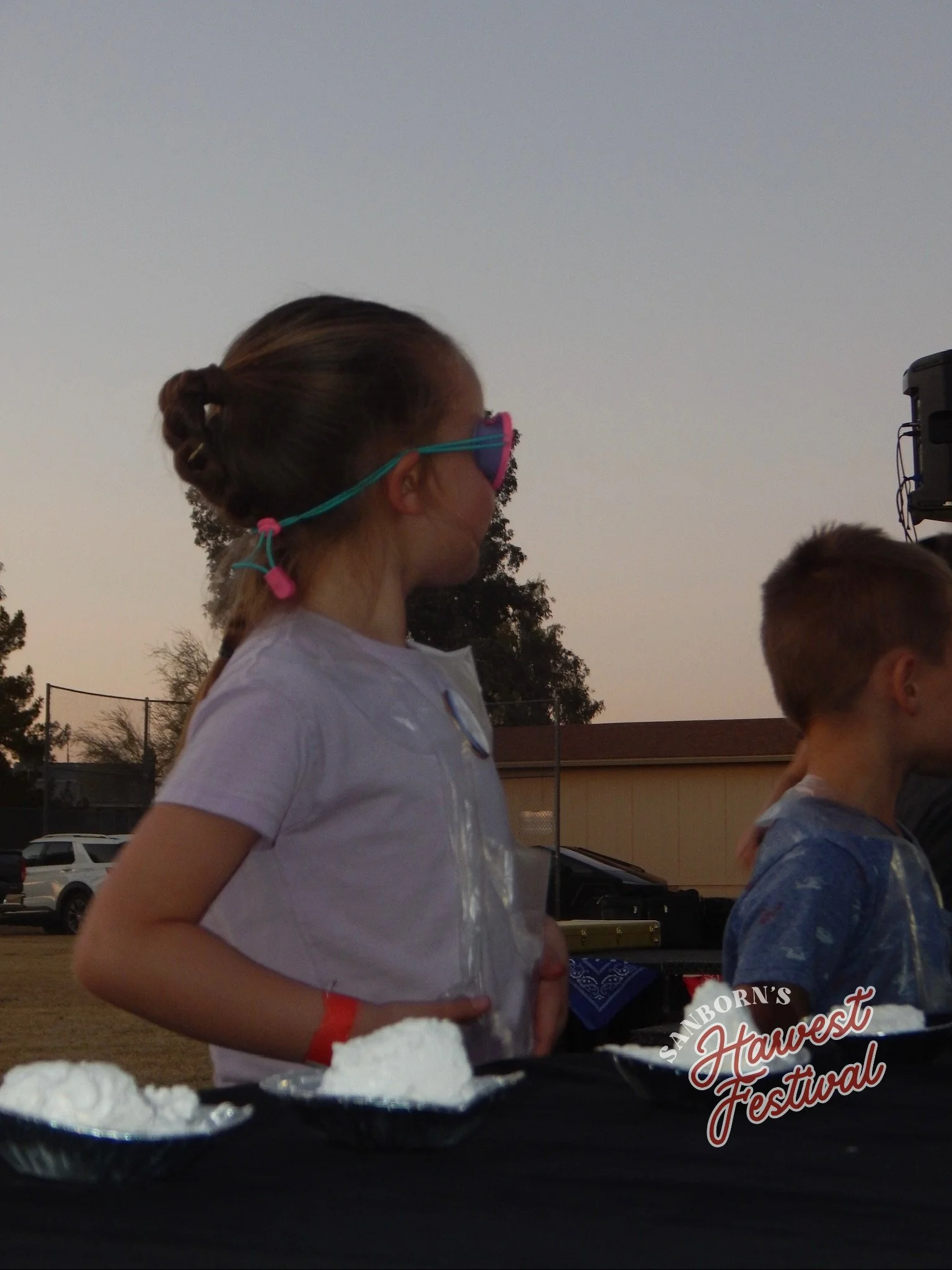 A girl wearing sunglasses and a white shirt stands outdoors at sunset during the Sanborn's Harvest Festival, with other people and a building in the background.