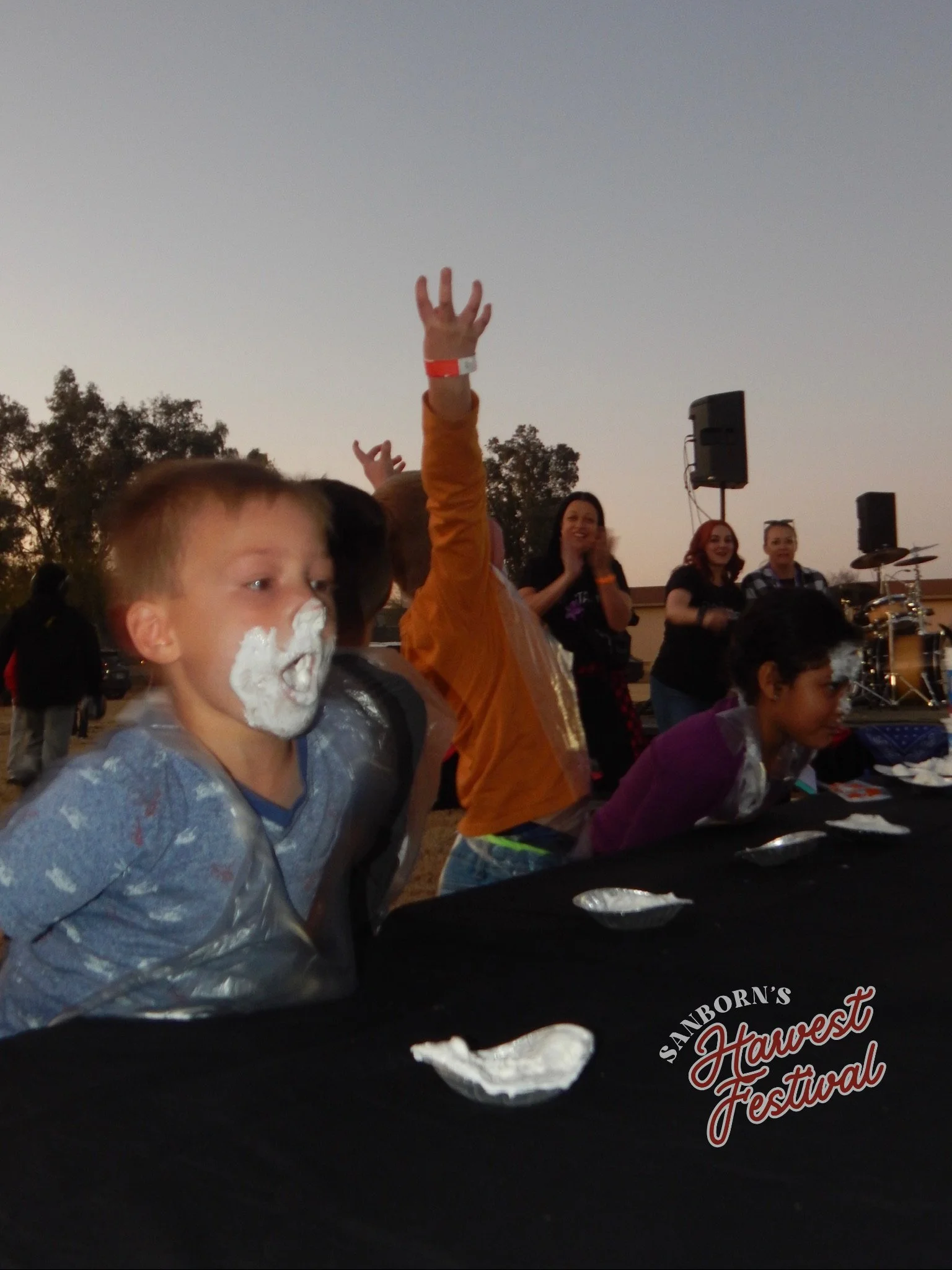 Children at the Sanborn's Harvest Festival eating pie, with a potluck-style setting in the evening. A group of women are seen clapping and smiling in the background, a musician is visible on a stage, and children are raising their hands, some with wh