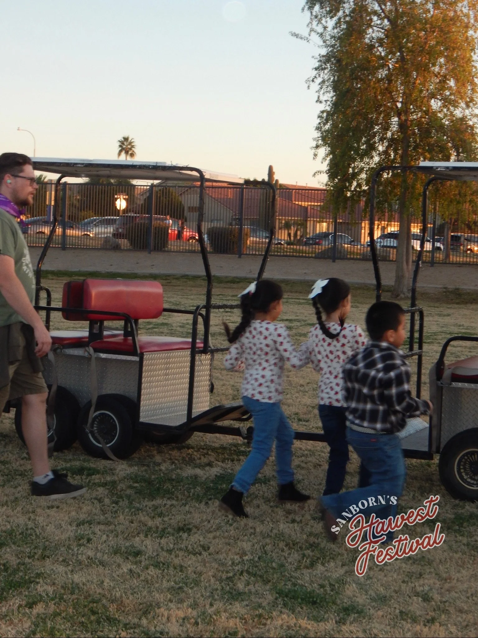 Children and an adult at a pumpkin festival with two hayride wagons on a grassy field during sunset. Trees and parked cars are in the background.