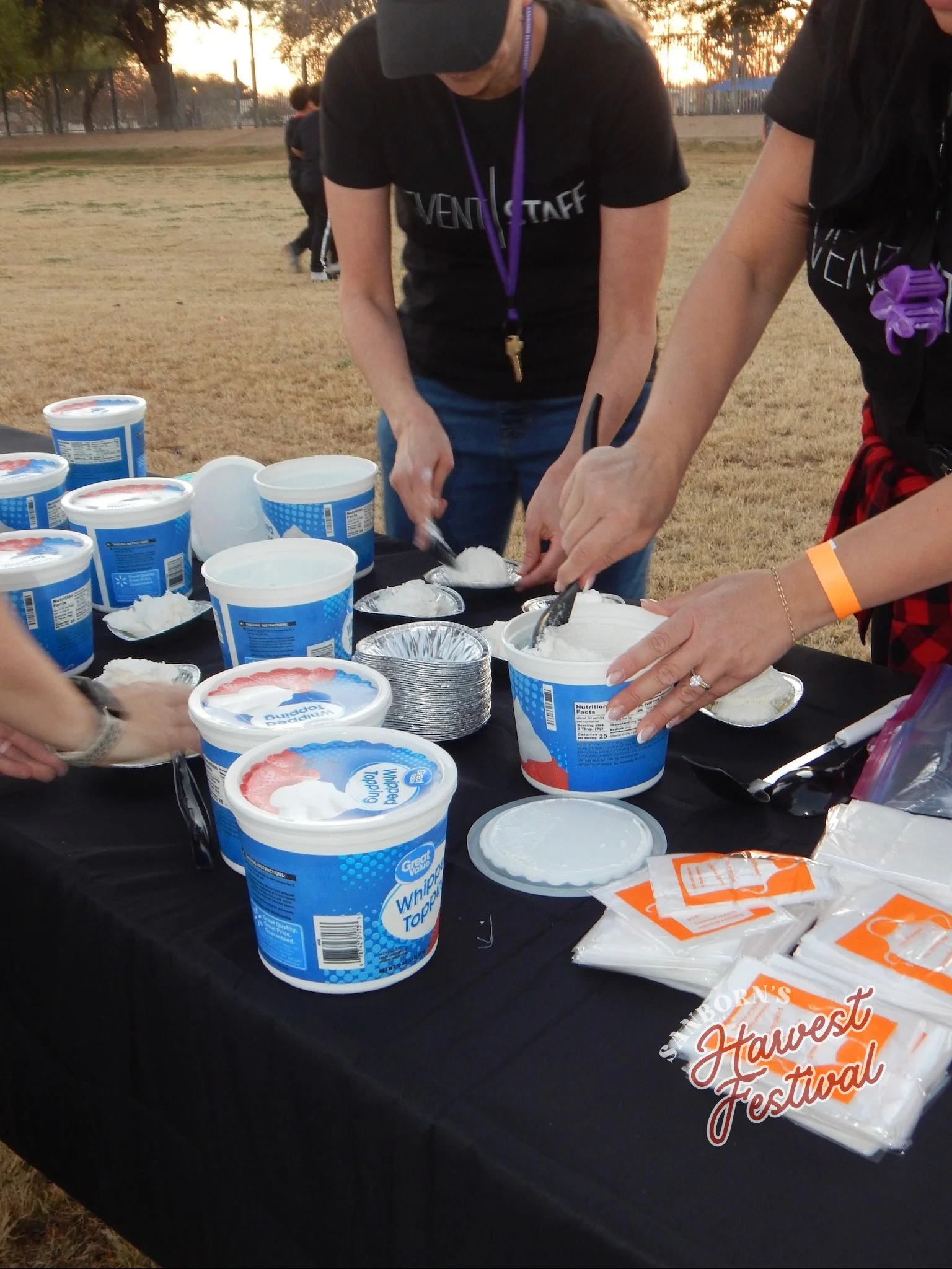 People preparing to serve or distribute whipped topping in containers at an outdoor event during sunset, with a black tablecloth and various food packaging on the table.