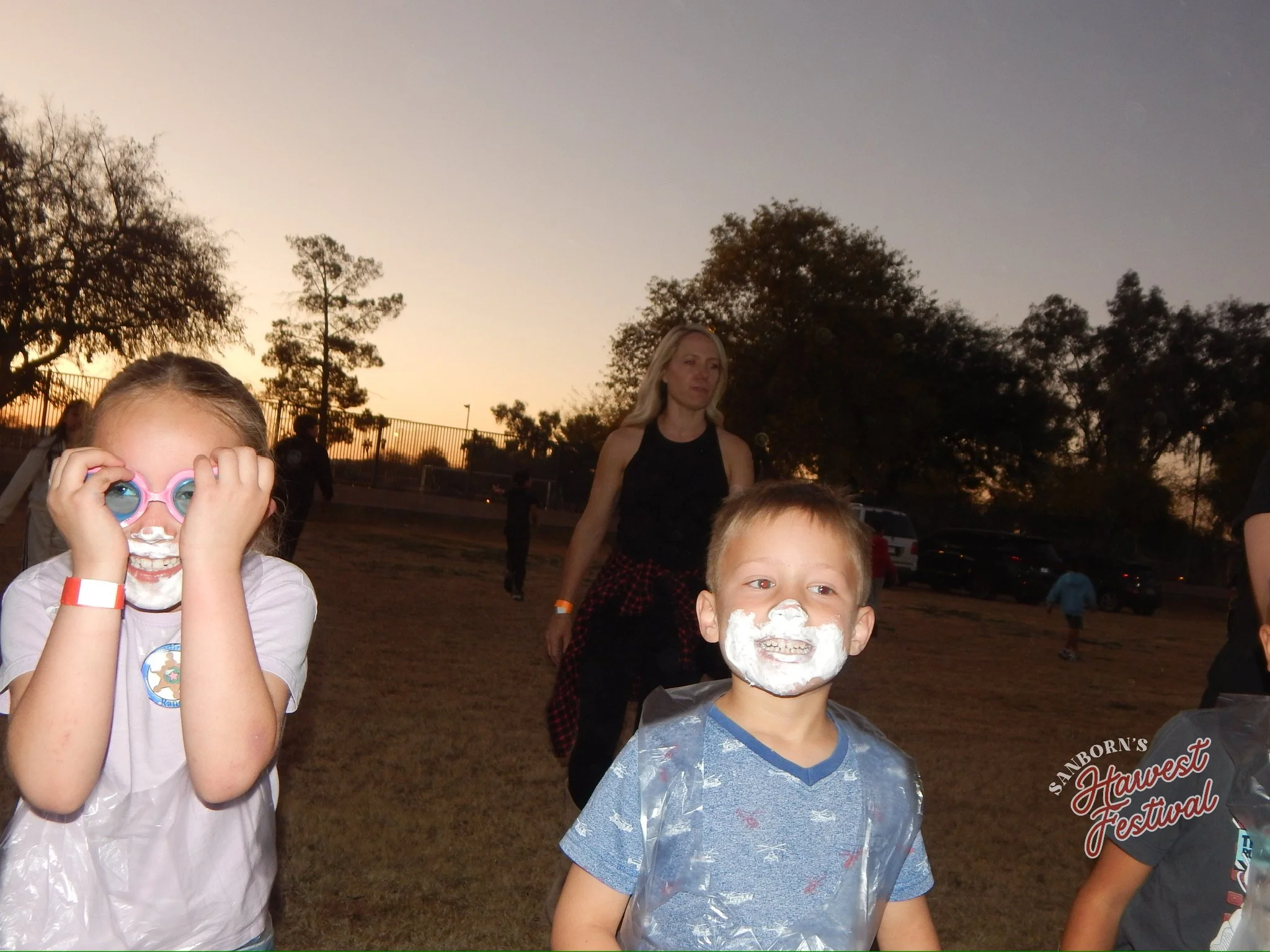 Children at an outdoor event during sunset with trees and a fence in the background. Two children are in the foreground, one girl with goggles smiling and a boy with face paint and a plastic apron, and a woman and other children are visible in the ba