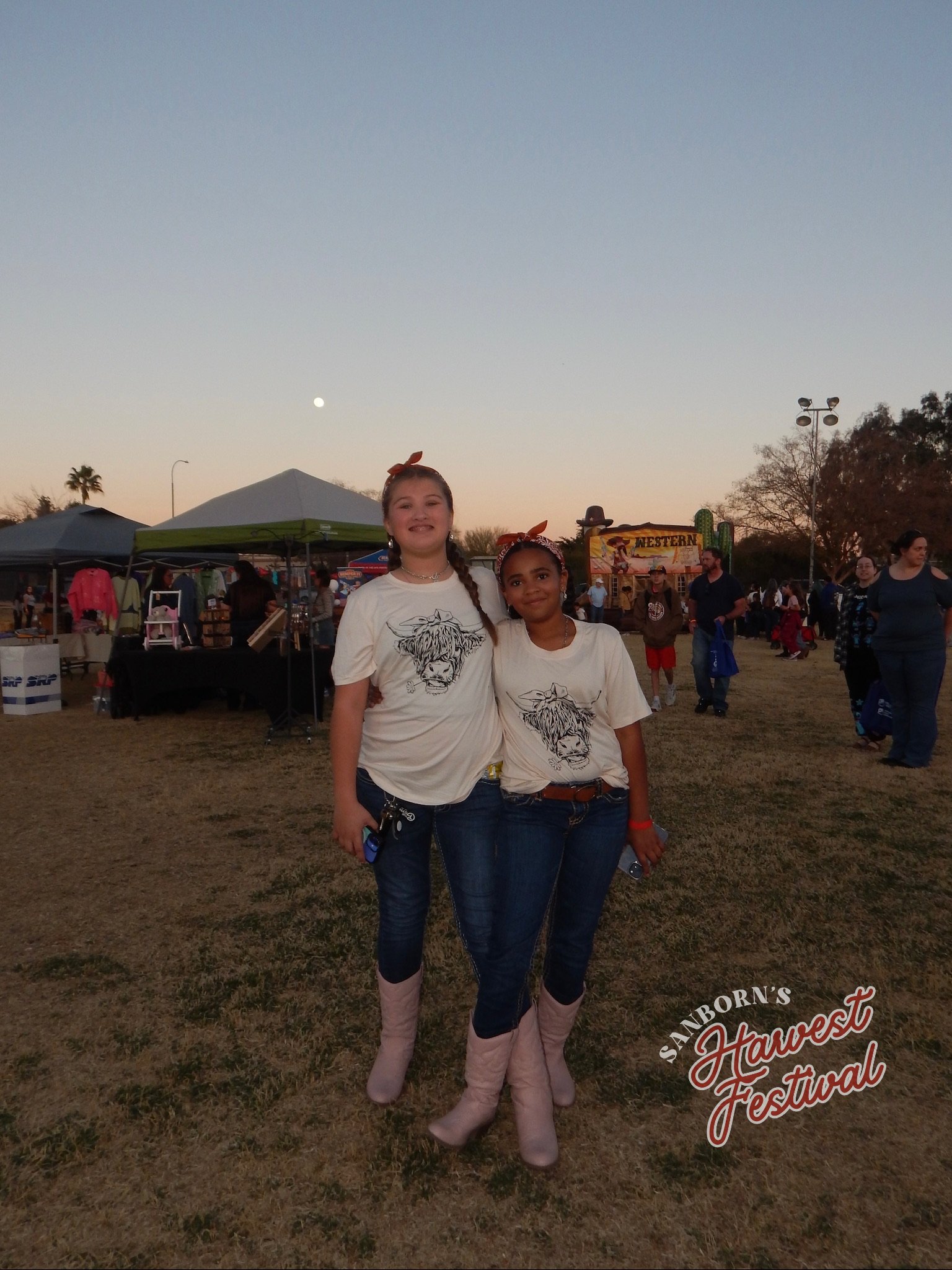 Two young girls standing together at an outdoor festival during sunset, wearing matching white t-shirts with a lion graphic, and pink cowboy boots, with vendor tents and festival signage in the background.