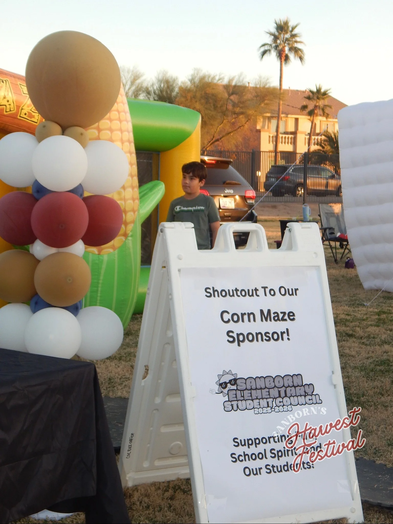 A boy standing behind a white sign at a school festival, with balloons and a large inflatable corn on the cob. The sign reads, 'Shoutout To Our Corn Maze Sponsor!' and lists the 'Sanborn Elementary Student Council 2025-2026' and 'Supporting School Sp