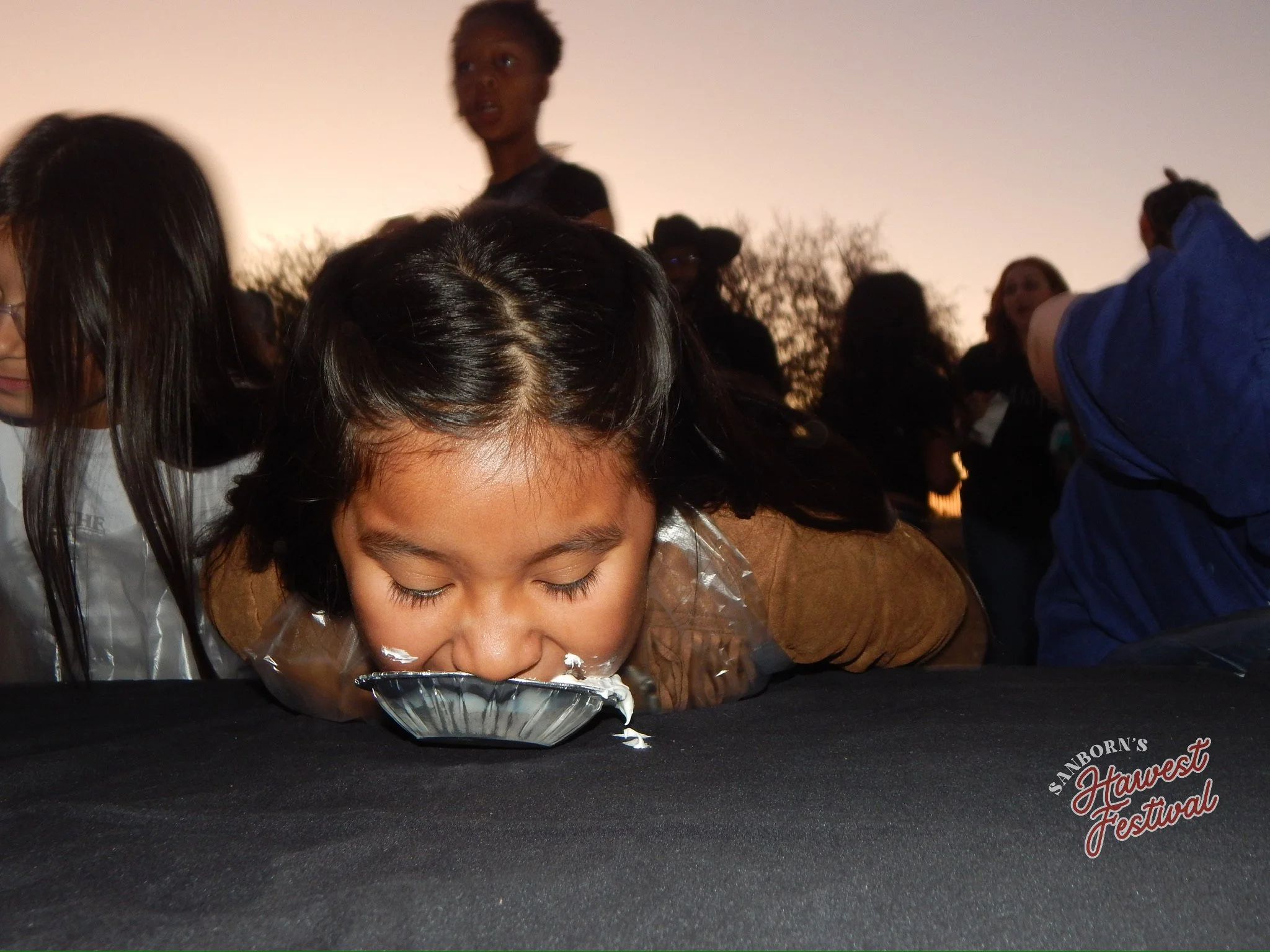 A young girl with dark hair leans over a black table, smelling or tasting a small silver-colored pie or dish at the Sanborn's Harvest Festival, with several people in the background during sunset.