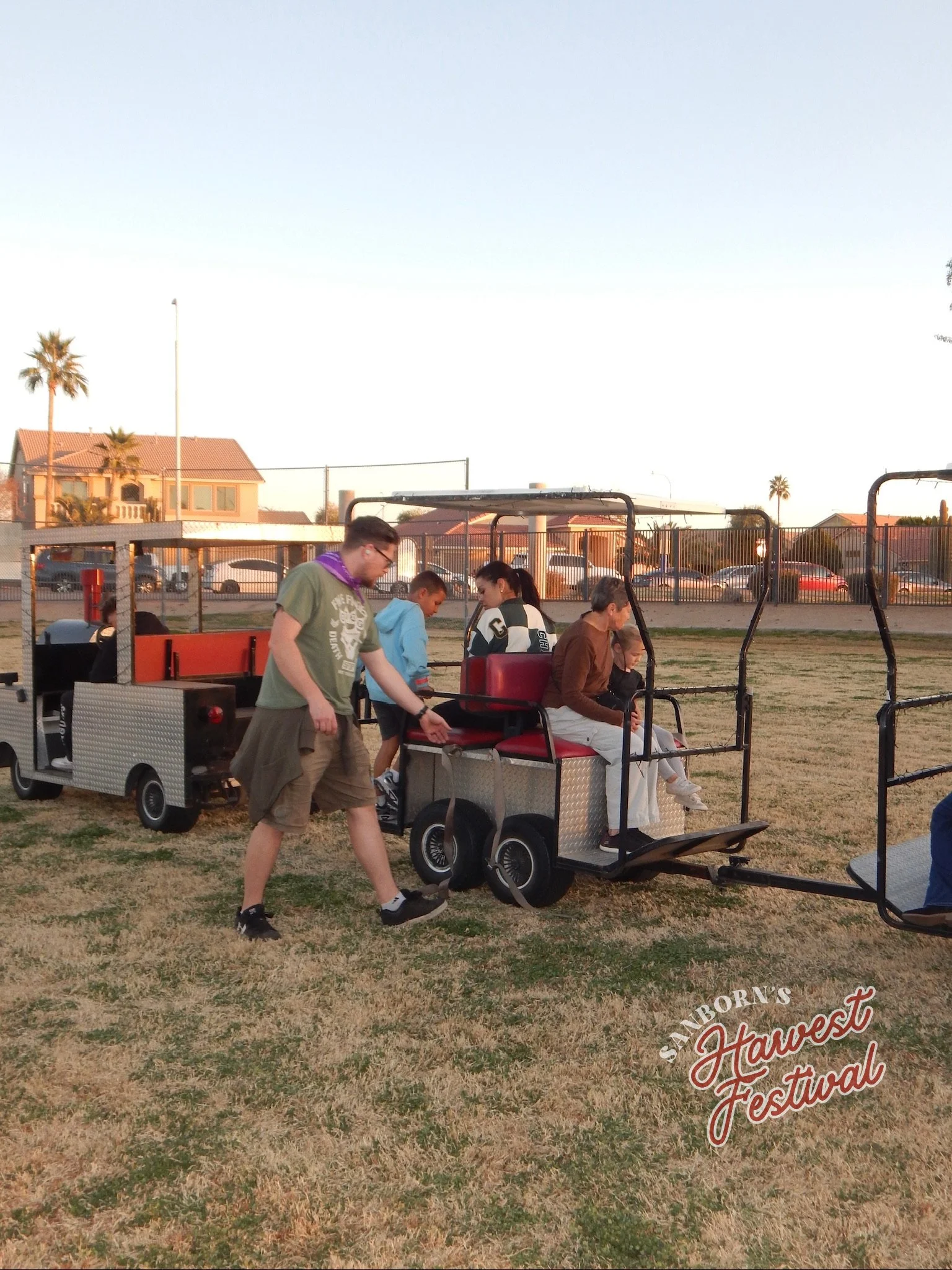 People riding a golf cart at the Sanborn's Harvest Festival in an outdoor park with houses and palm trees in the background.