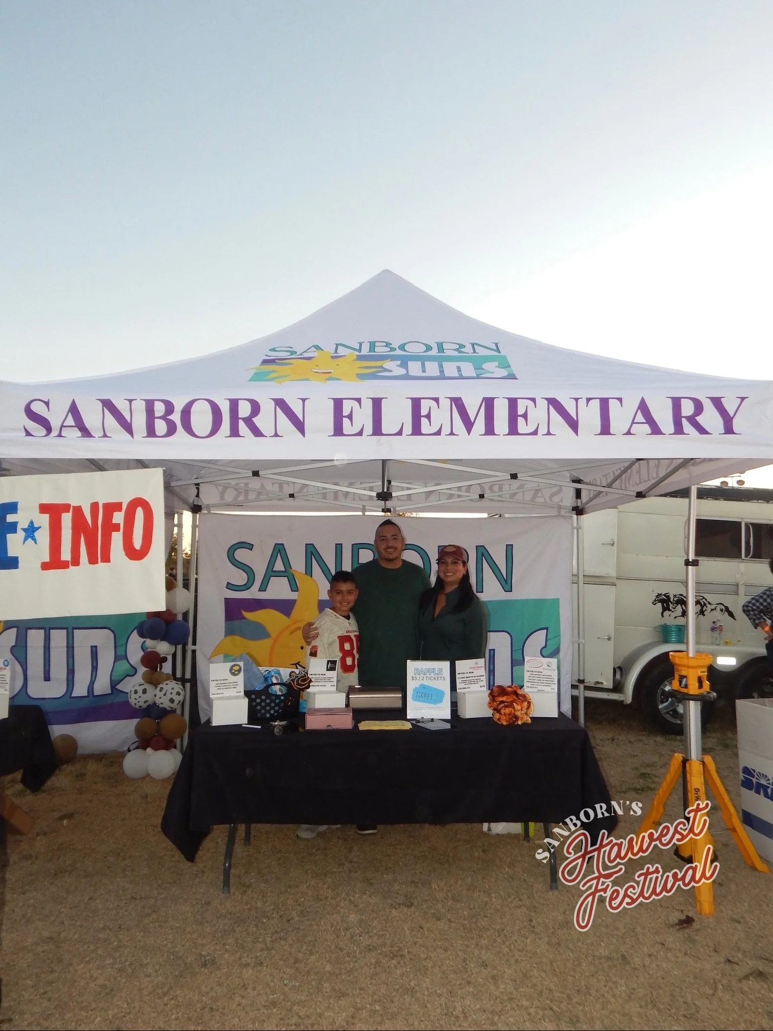 A man, woman, and boy standing inside a tent at the Sanborn Elementary School Harvest Festival, with raffle tickets and promotional items on a table in front of them.