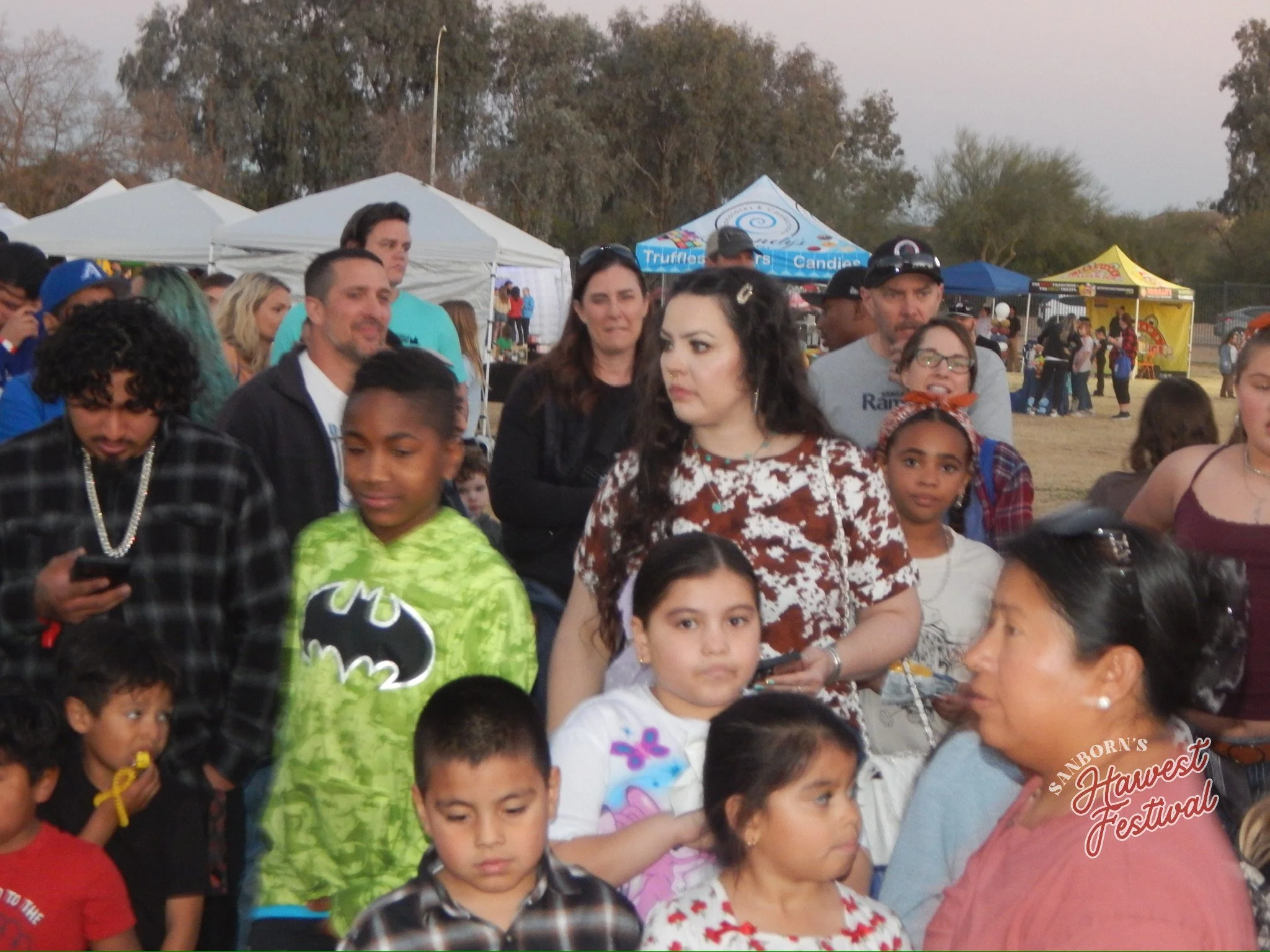 A large group of people gathered outdoors at a festival with white and colorful tents in the background. Many individuals are looking in various directions, some using their phones, with children and adults present. The overall atmosphere appears liv