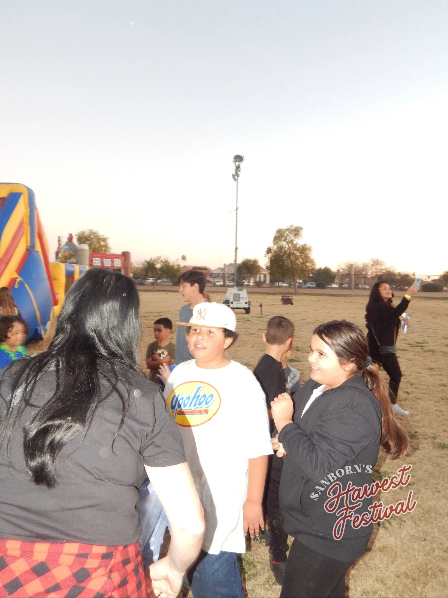 Group of people at an outdoor festival, some are smiling, talking, and taking photos. There are trees, a yellow and blue inflatable structure, and a sign that says 'Sanborn's Harvest Festival' in the background.