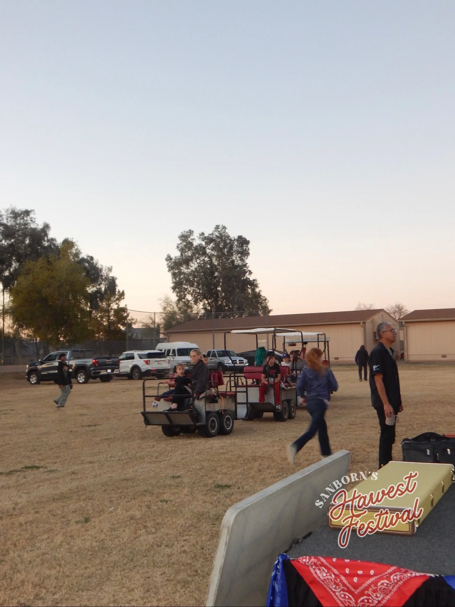 People gathering at the Sanborn's Harvest Festival during the evening, with a ride-on trailer carrying children, trees, parked cars, and a building in the background.