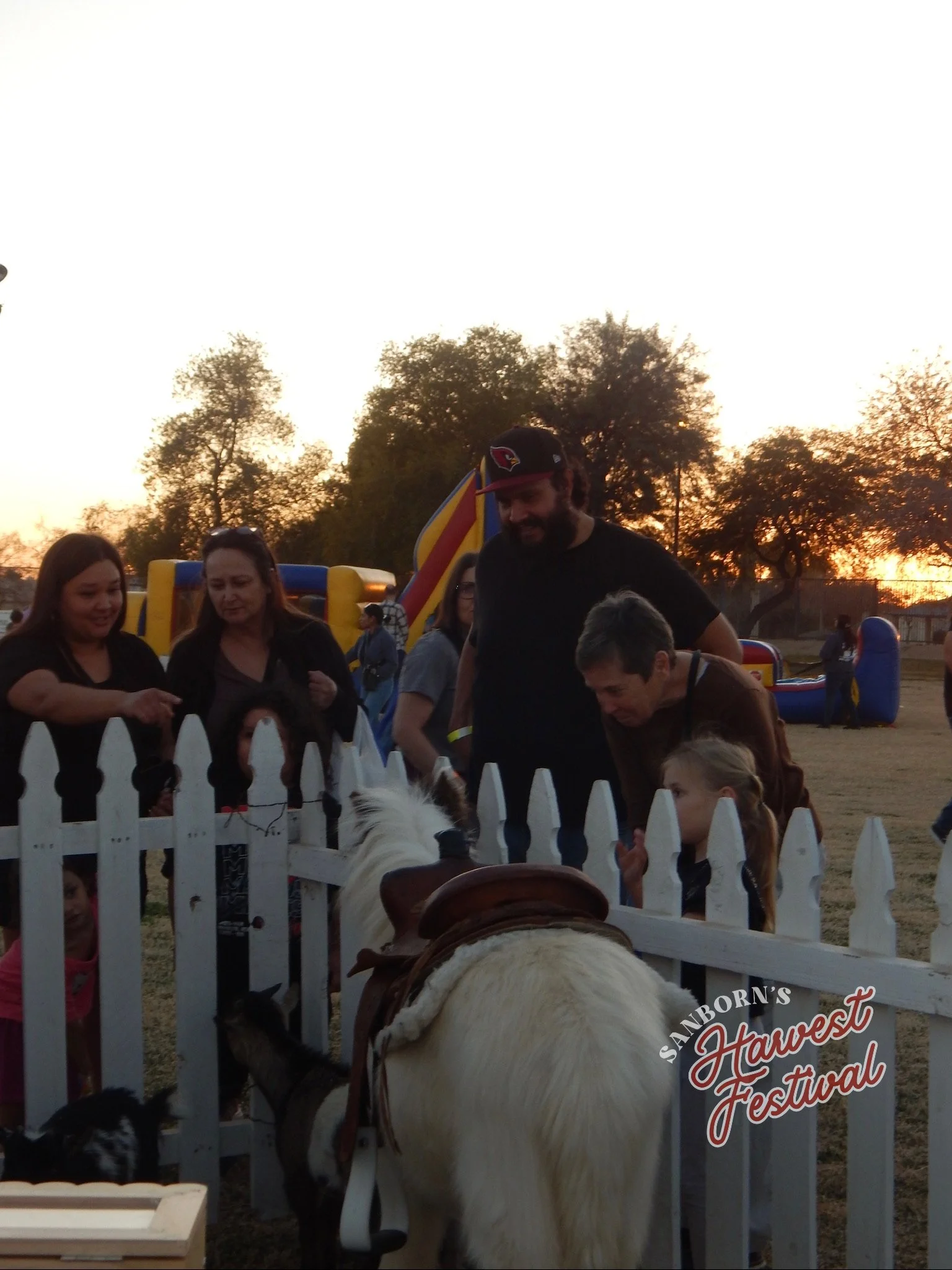 People gathered around a white picket fence at sunset during the Sanborn's Harvest Festival, with trees and inflatables in the background.