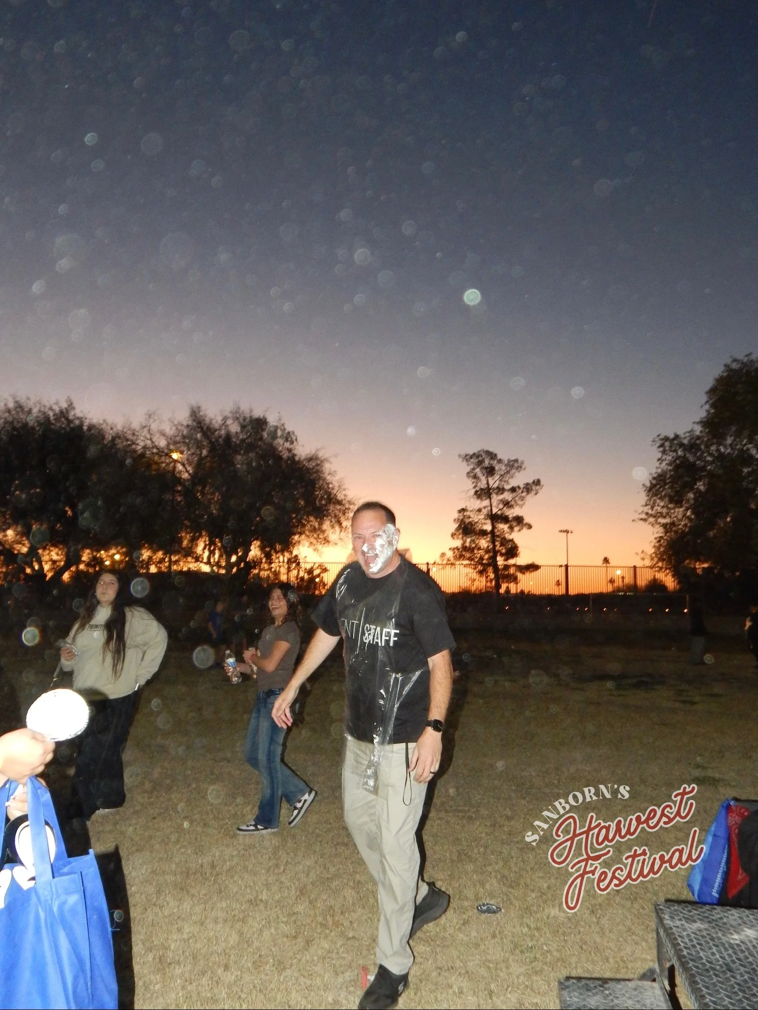 Person with face covered in white foam at sunset during an outdoor festival, with other people around and trees in the background.