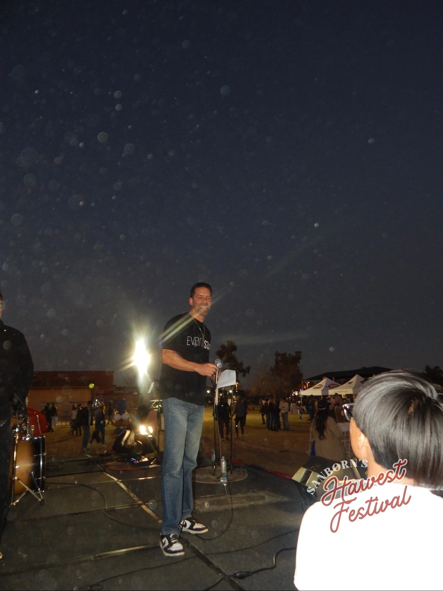 A man standing with a microphone in front of a crowd at night during an outdoor event or festival with tents and people in the background.