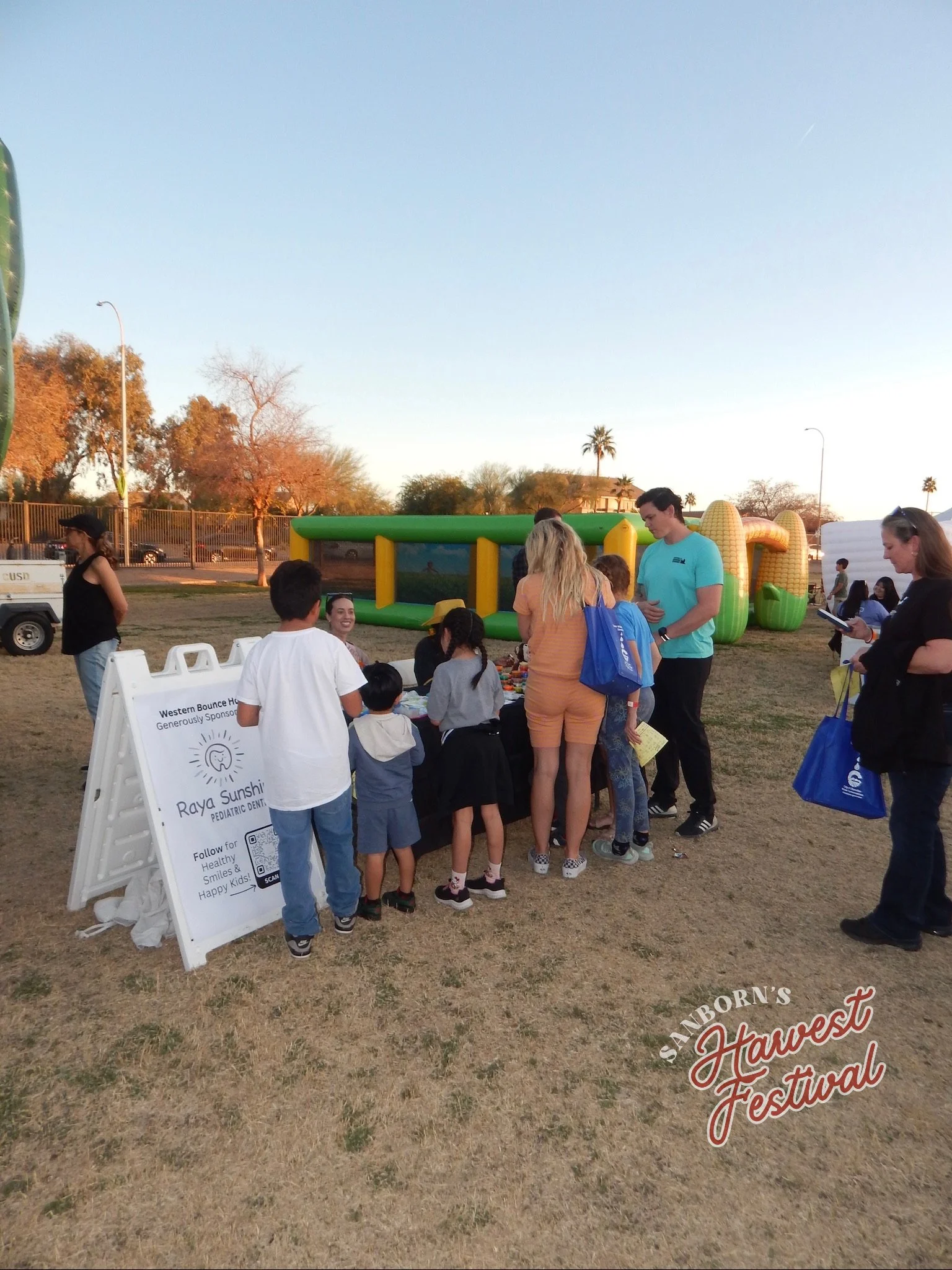 People gathered at a community event near inflatable play structures with trees and a fence in the background, during late afternoon or early evening.