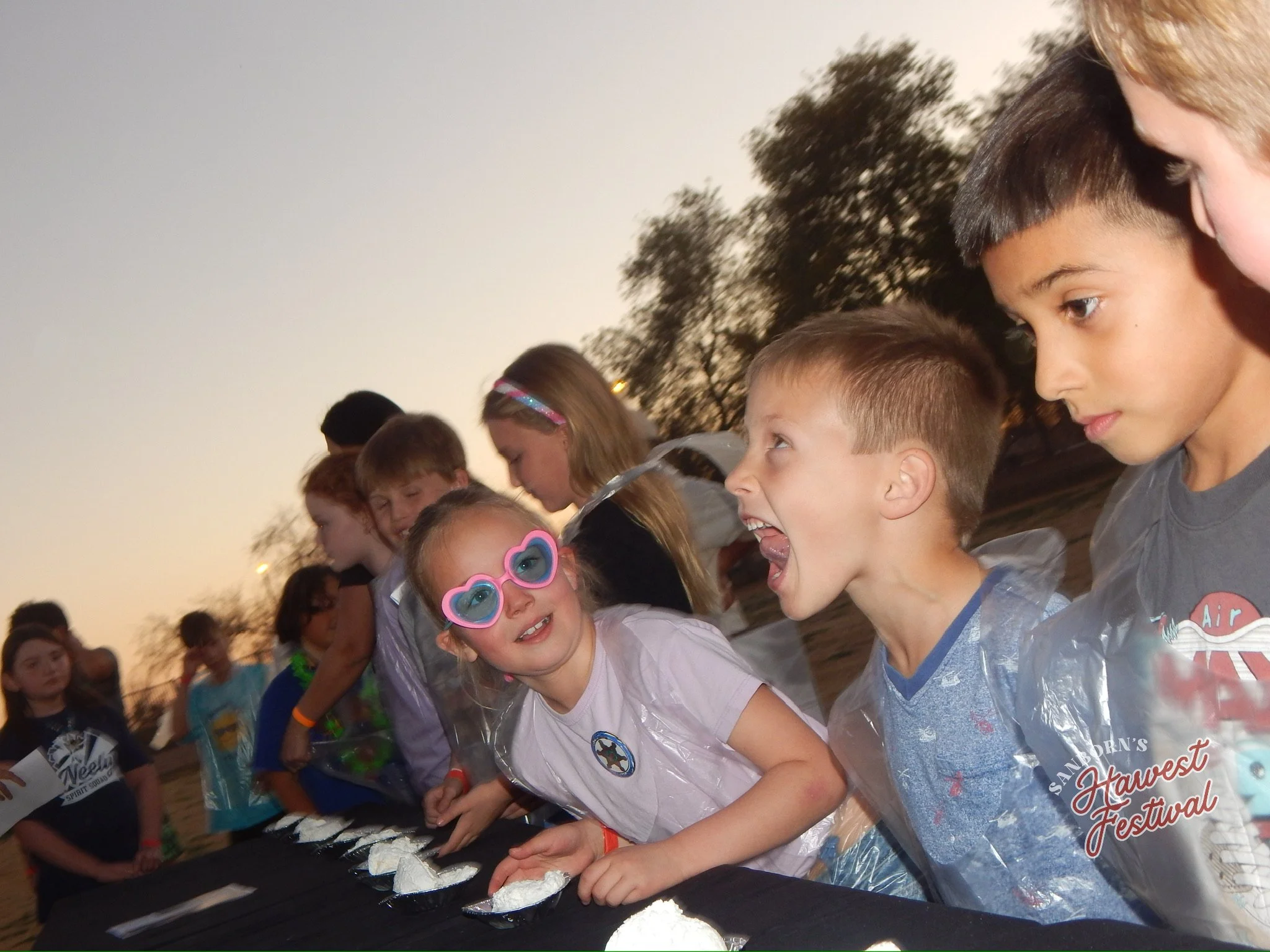 Children gathered at a festival, wearing casual clothes and colorful sunglasses, participating in an activity together, with trees and a sky in the background.