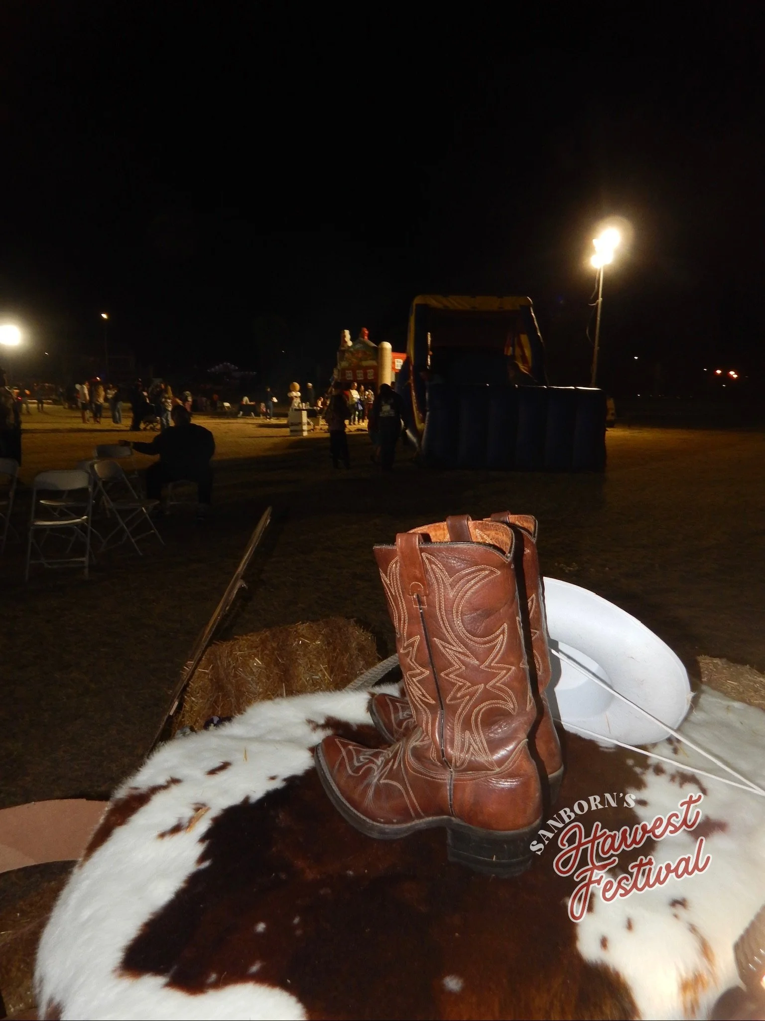 Night scene at a festival with people gathered, lights illuminating the area, and a rodeo bull in the foreground with a cowboy boot and a white cowboy hat resting on it. The event is called 'Sanborn's Harvest Festival' as seen on the logo.