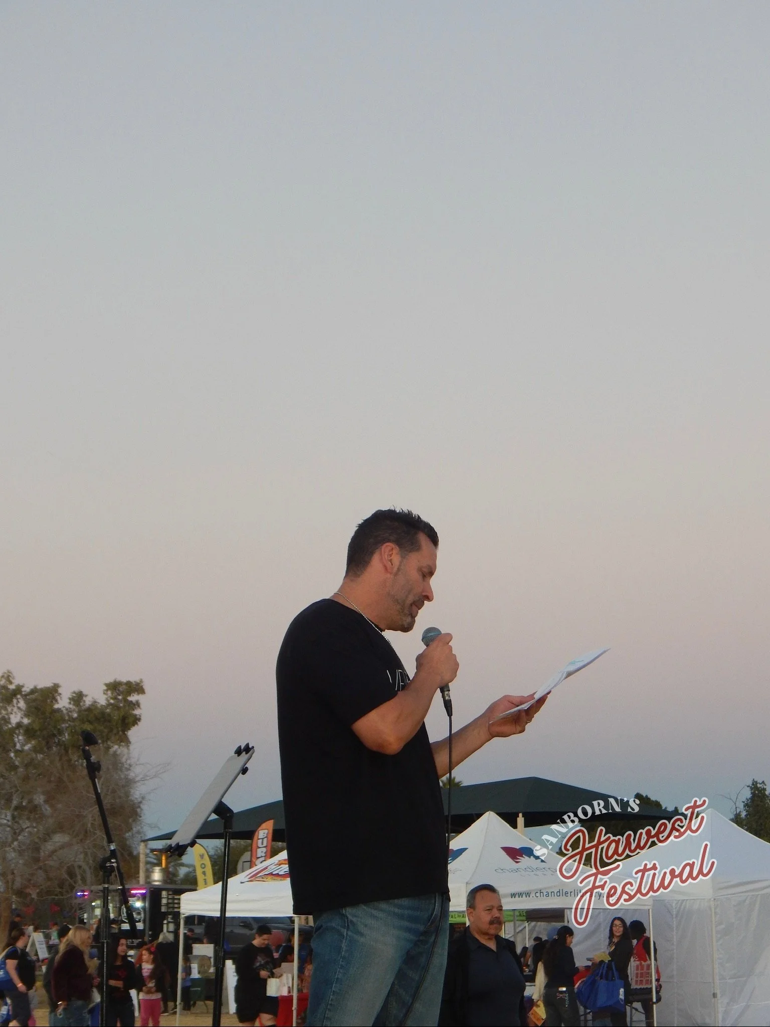 A man holding a microphone and reading from a paper at an outdoor festival with tents and people in the background.