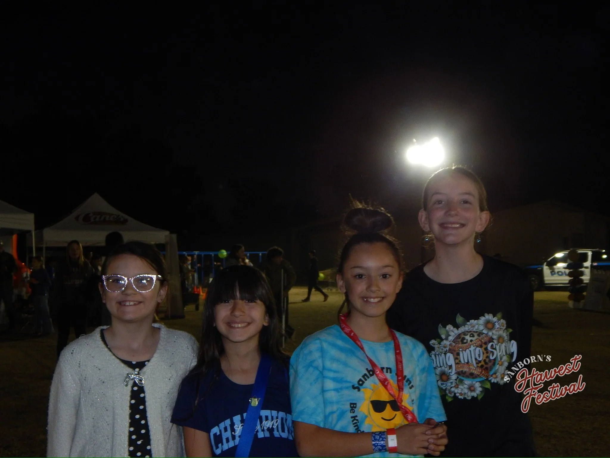 Four smiling young girls at a nighttime outdoor festival, dressed casually, standing close together, with tents, lights, and a police car visible in the background.